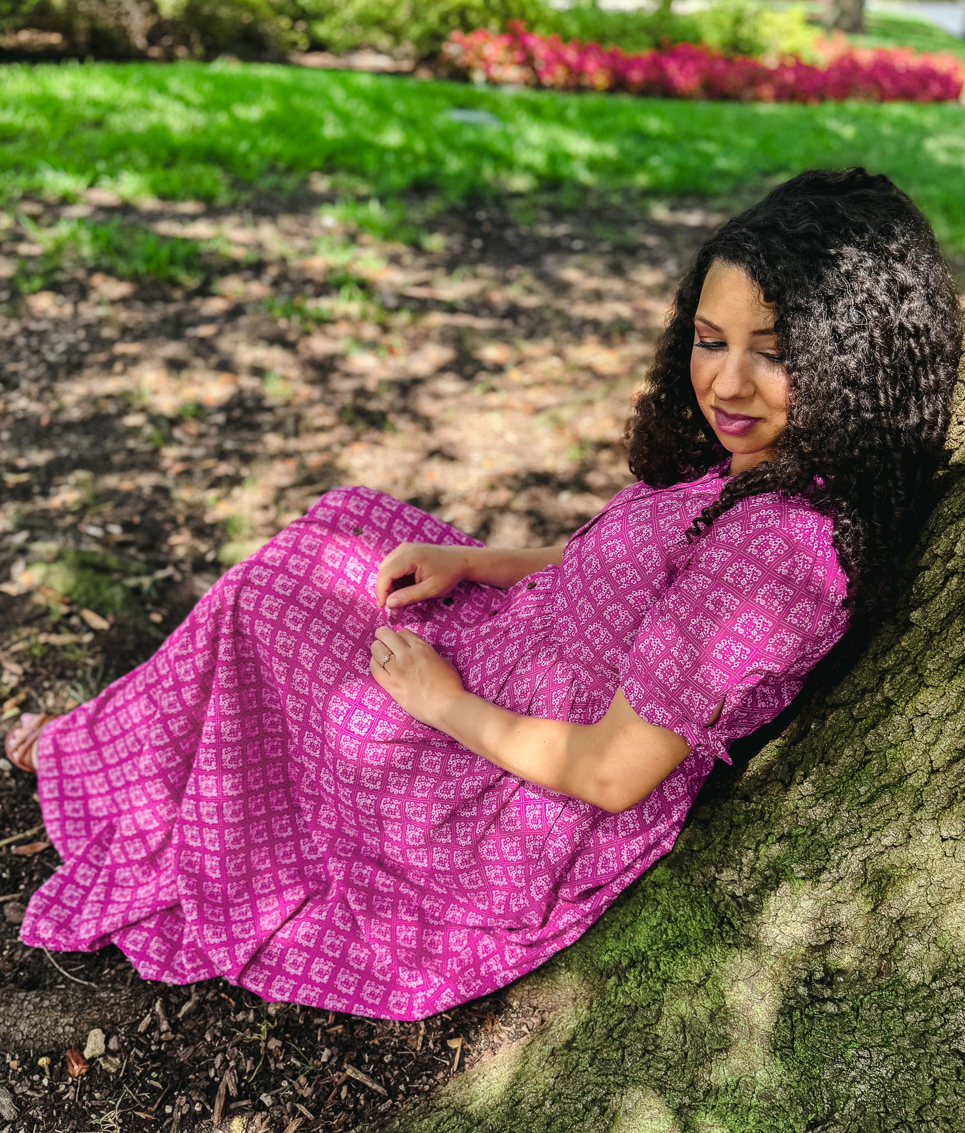 Woman in a pink modest nursing dress sitting against a tree in a park