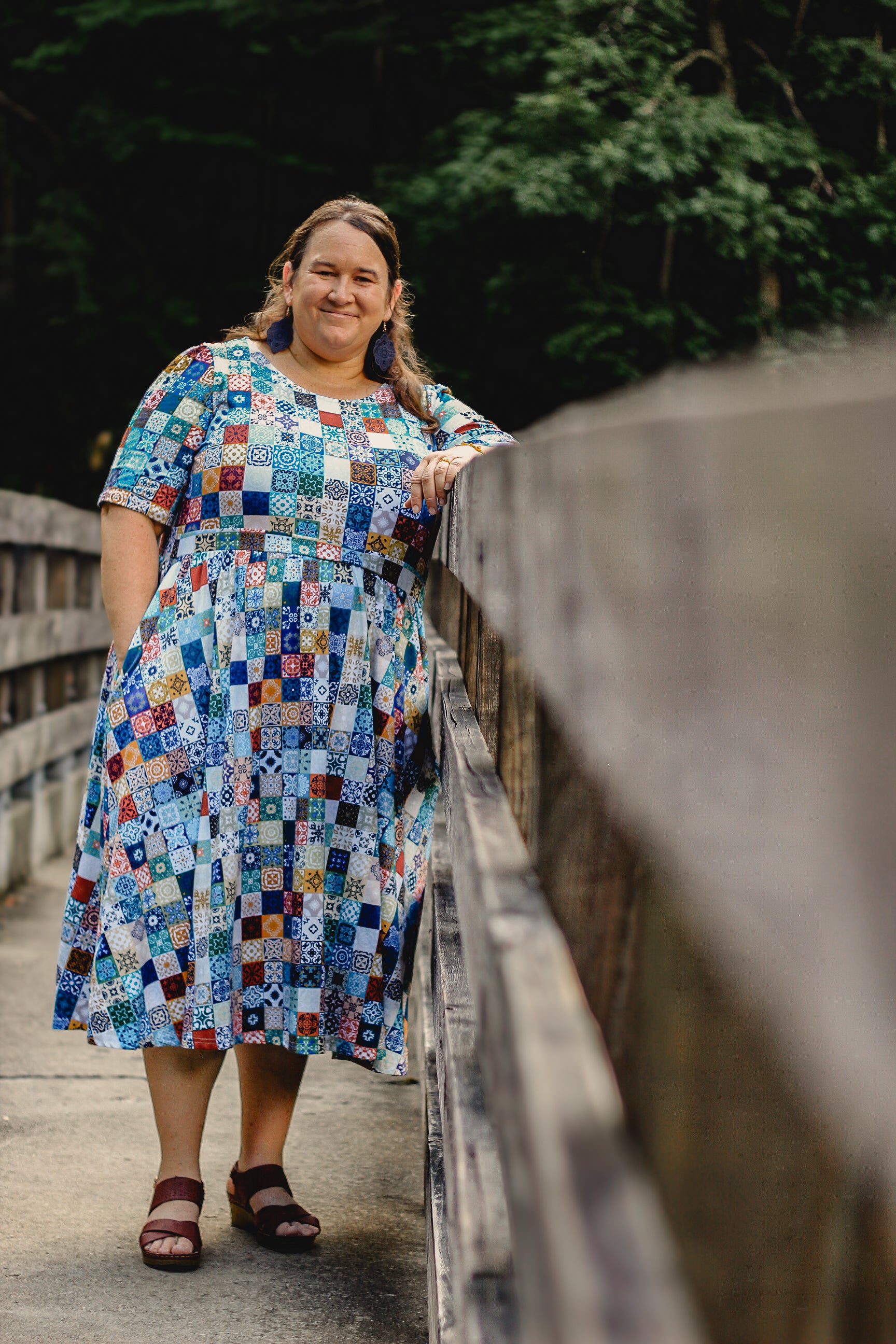 Woman in a colorful modest nursing dress standing on a wooden bridge with greenery in the background