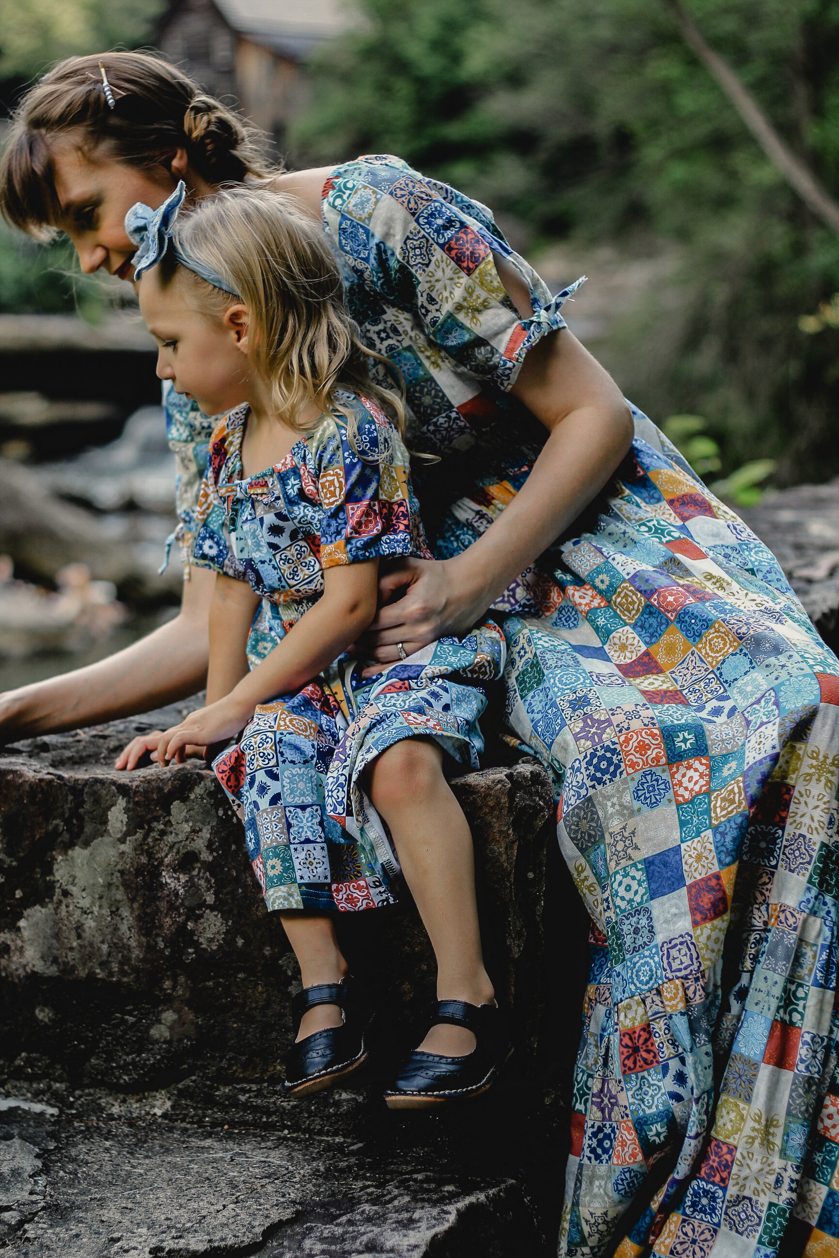 Mother and child in colorful floral modest dresses sitting on a stone ledge outdoors.