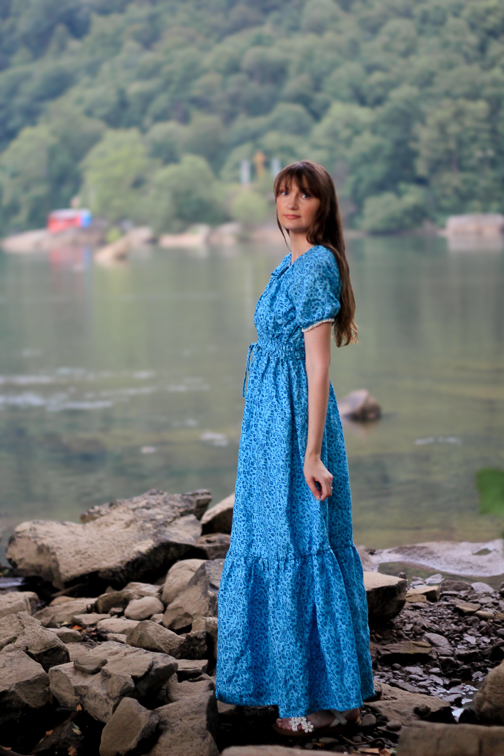 Woman in a blue modest nursing dress standing by a lake with trees in the background