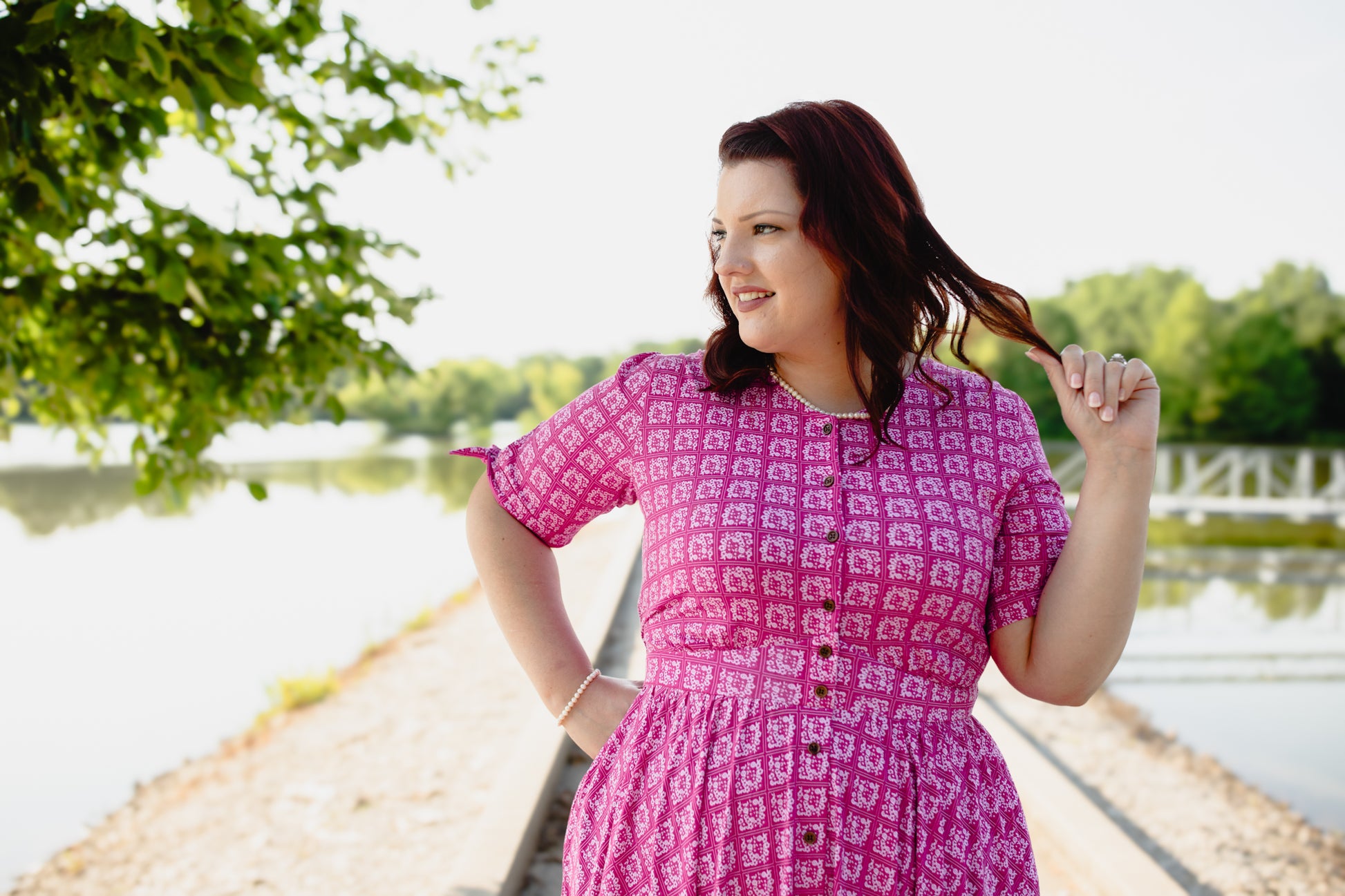 Woman in a pink modest nursing dress standing by a lakeside with trees in the background