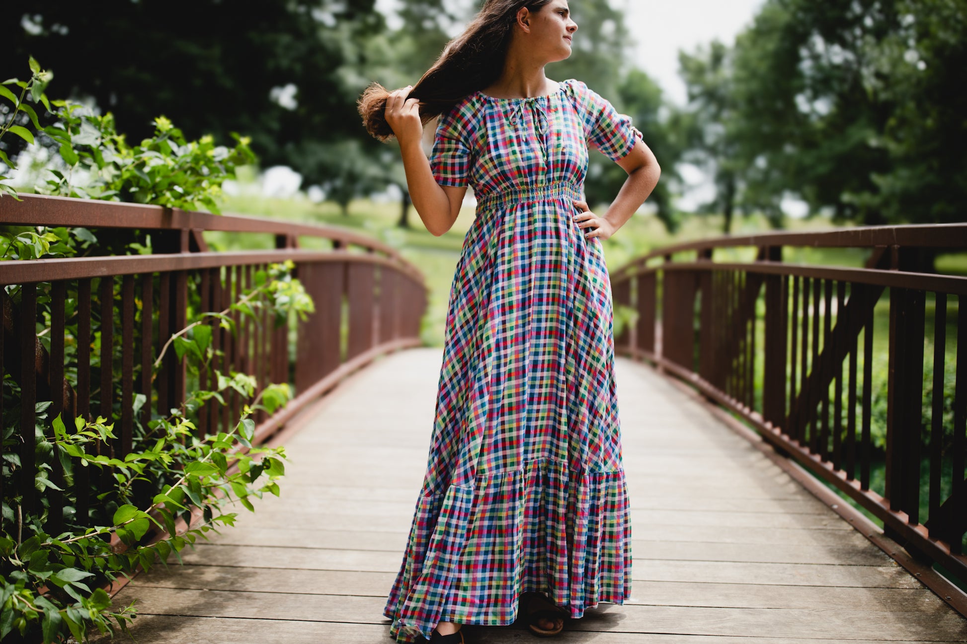 Woman in a plaid modest nursing dress standing on a wooden bridge with greenery around
