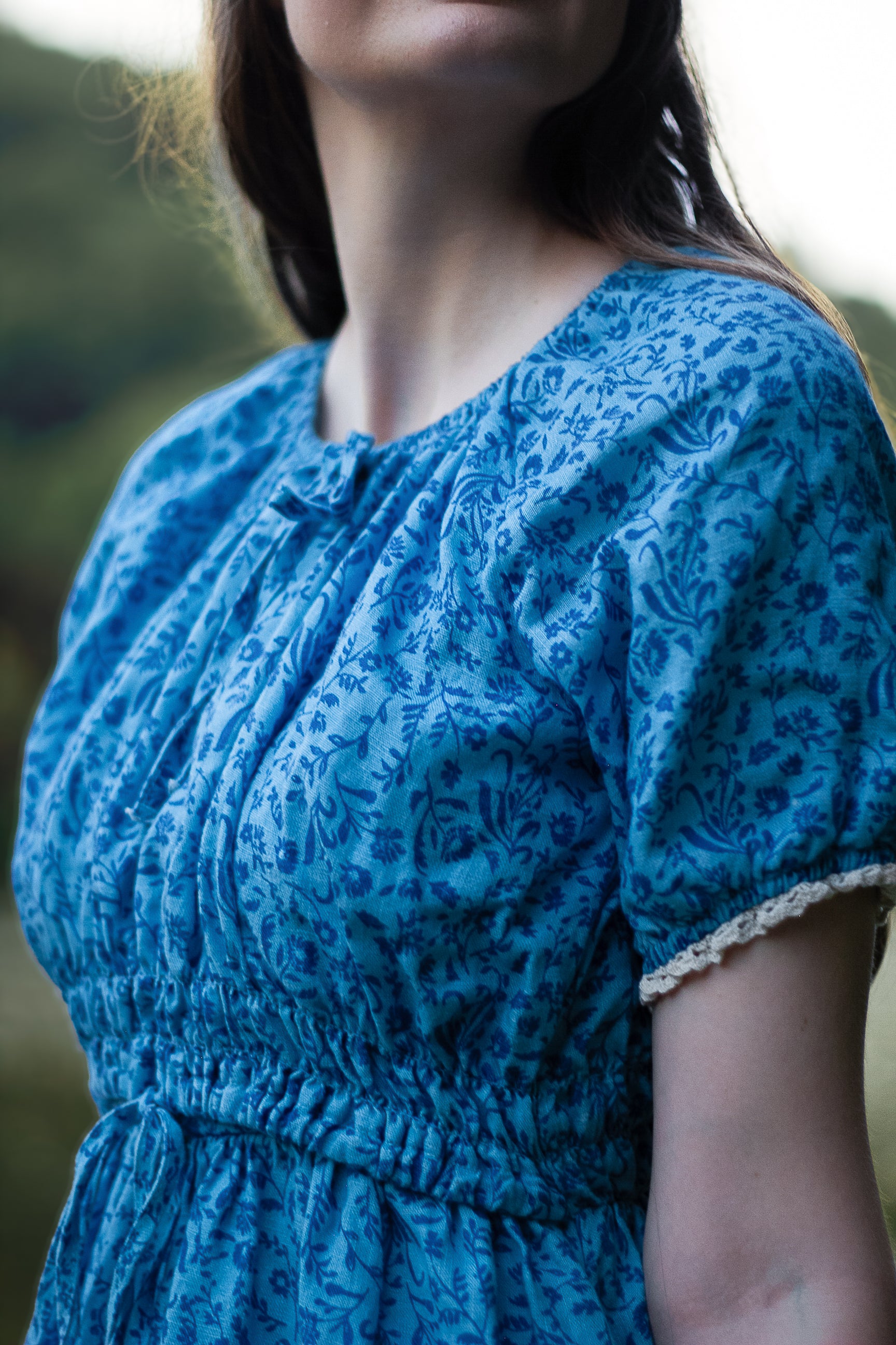 Woman wearing a blue modest nursing dress with a floral pattern against a blurred natural background
