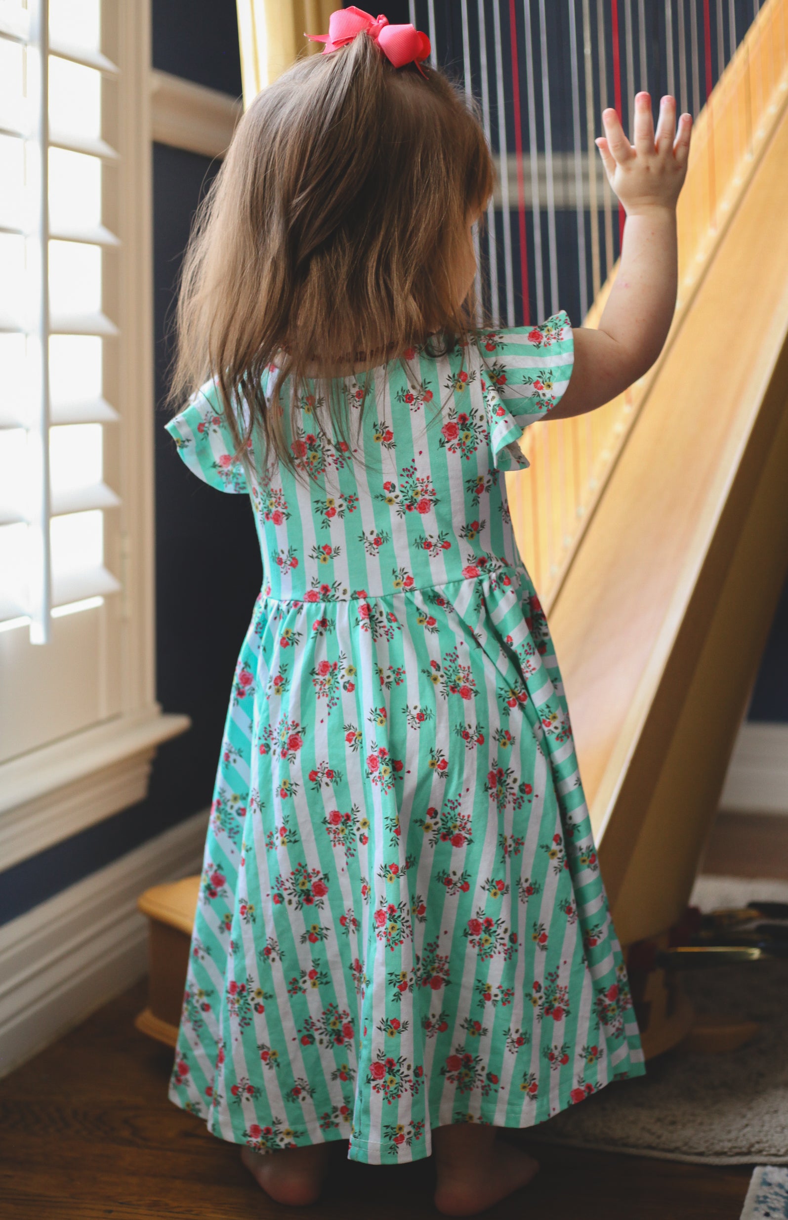 Child in a floral modest dress standing near a harp indoors.