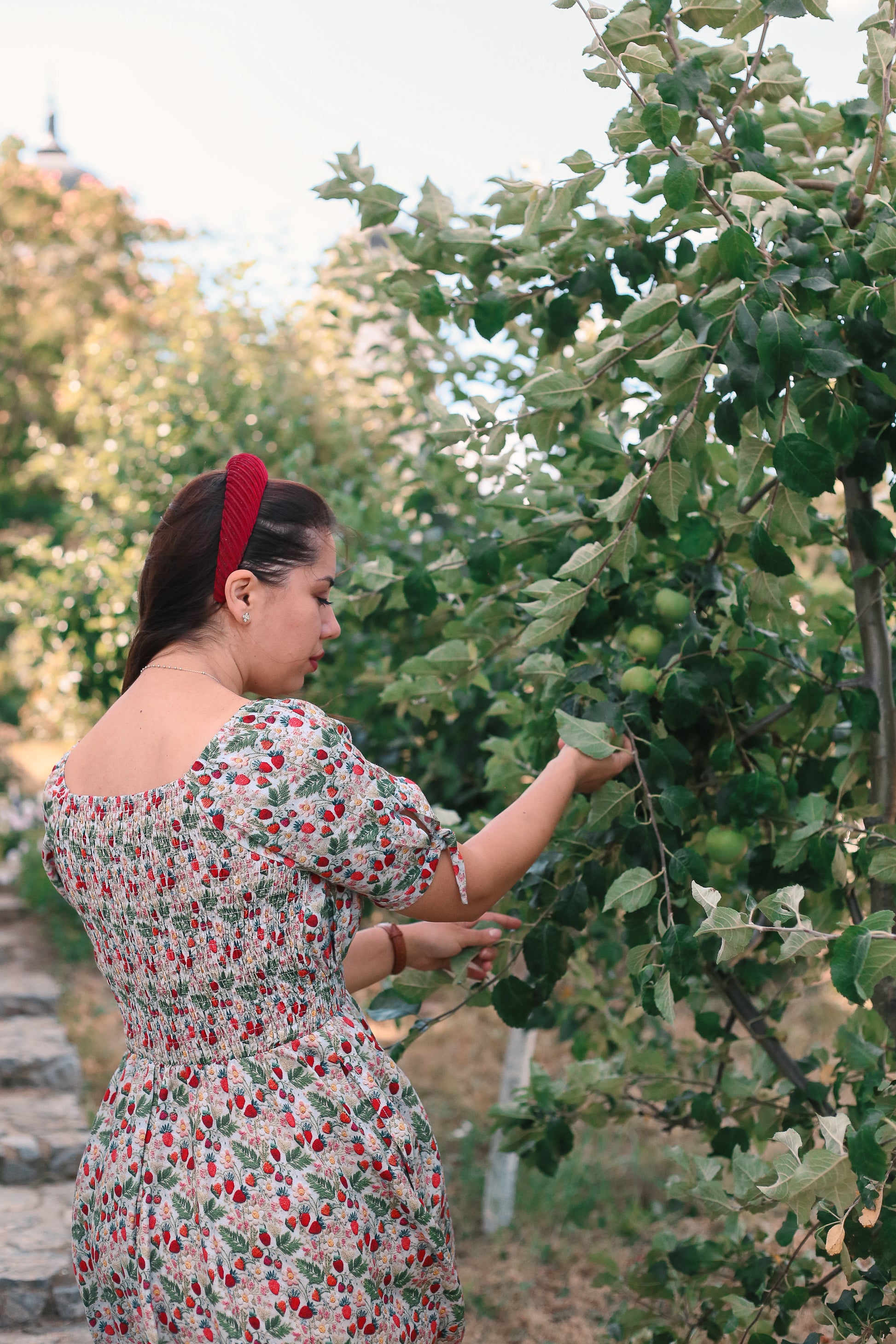 Woman in a floral modest nursing dress examining green apples on a tree