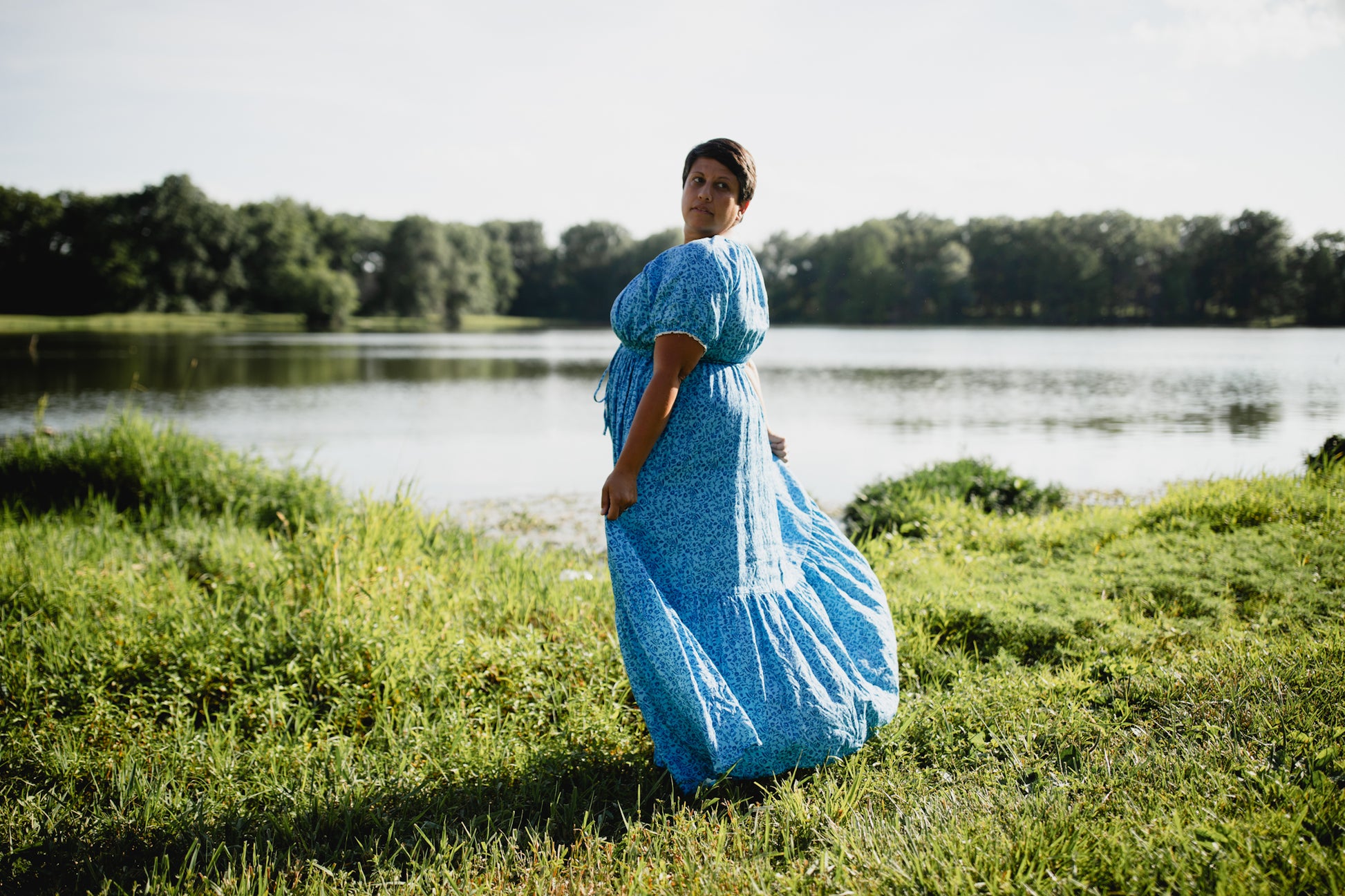 Woman in a blue modest nursing dress standing in a grassy area near a lake with trees in the background.