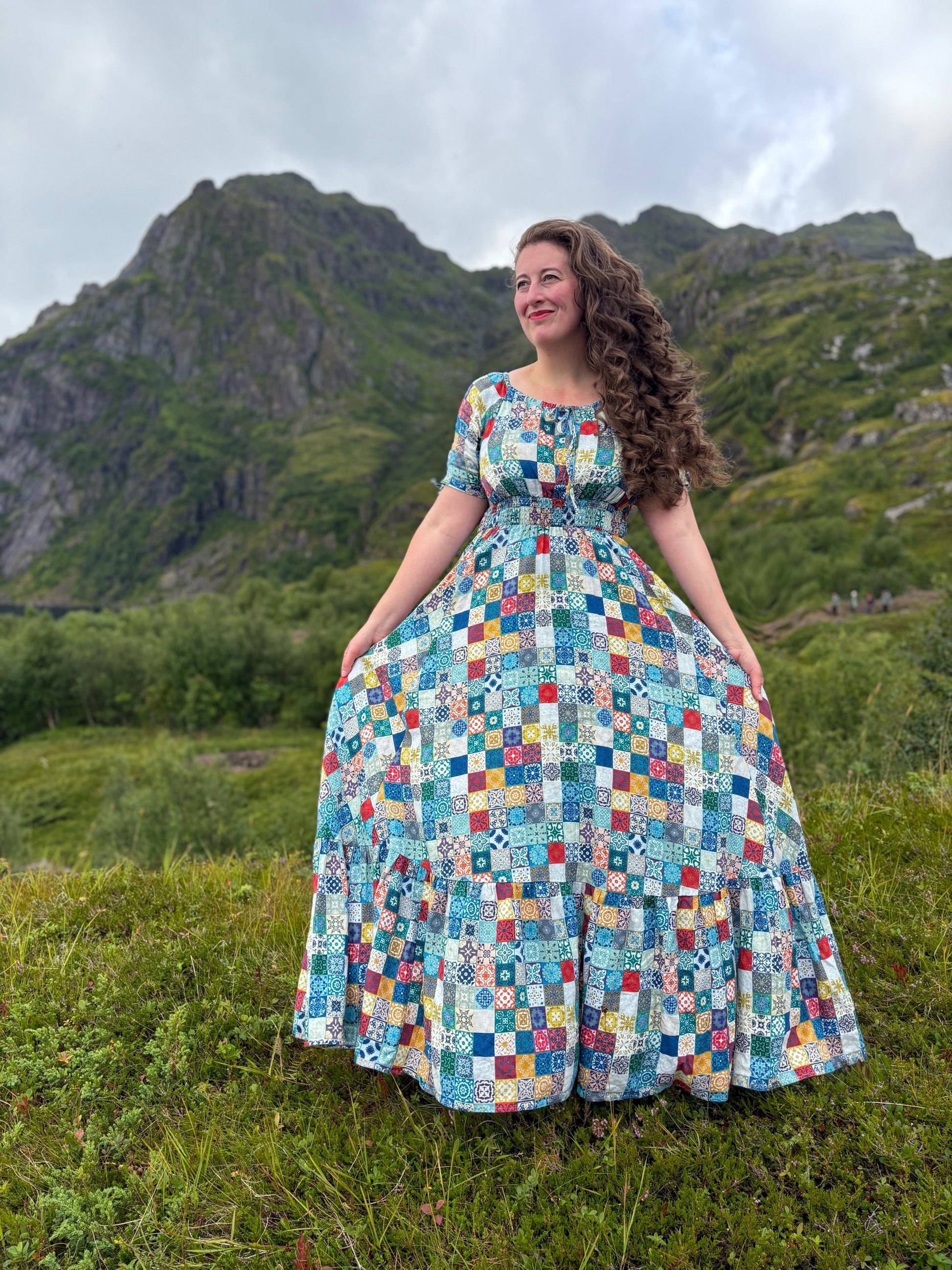 Woman in a colorful modest nursing dress standing in a grassy field with mountains in the background