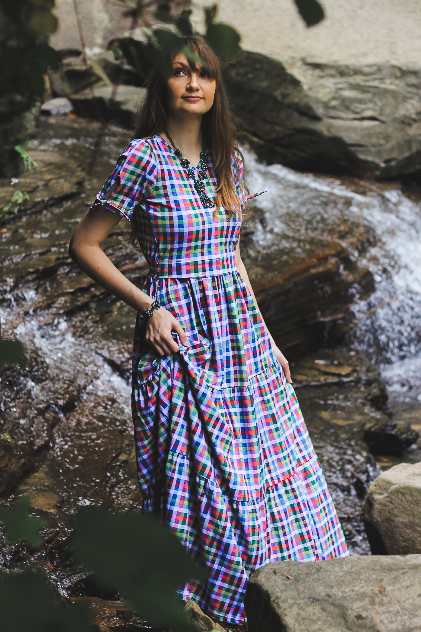 Woman in a colorful modest nursing dress standing by a stream with rocks and waterfalls in the background
