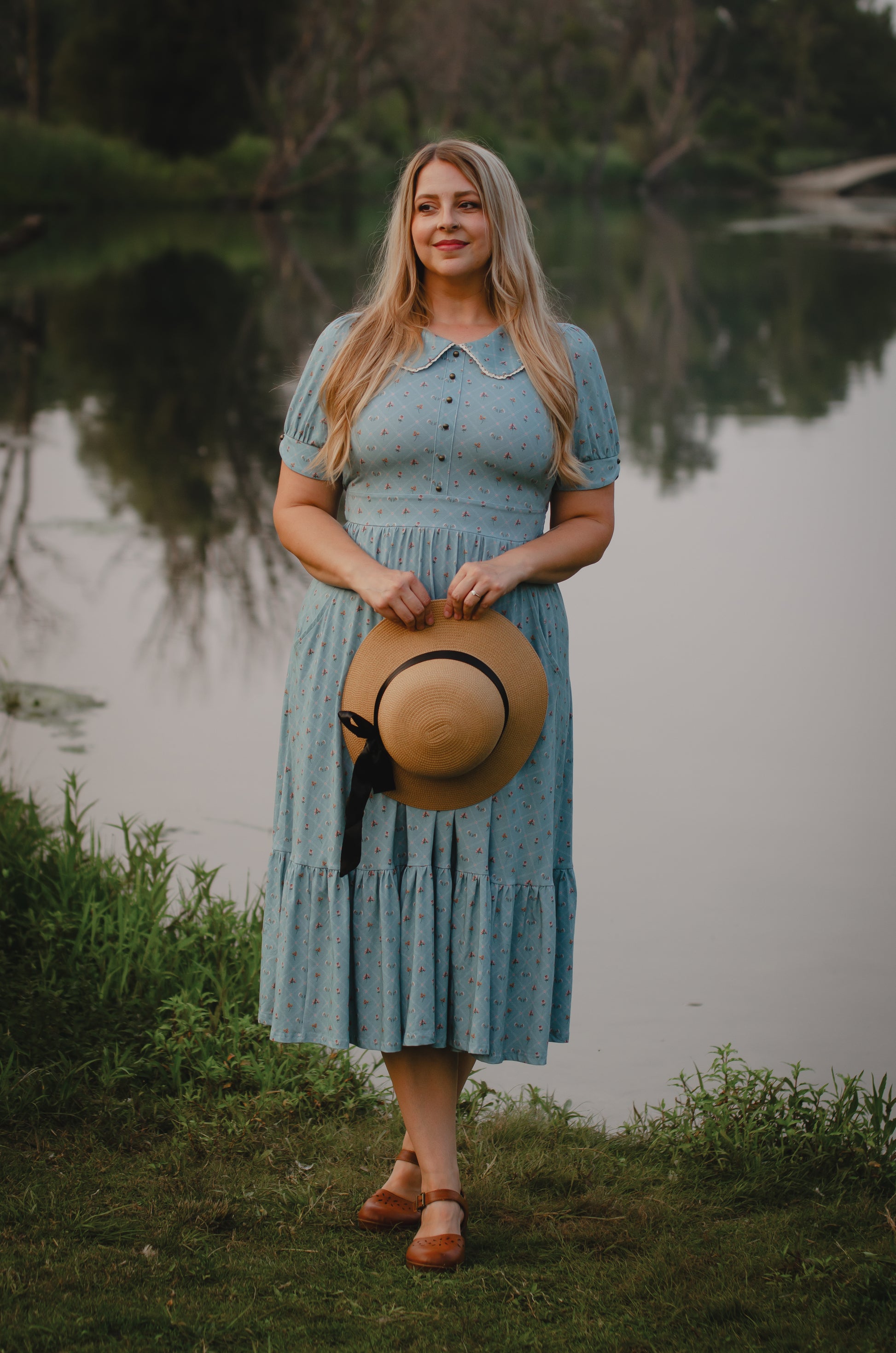 Woman in a blue modest nursing dress holding a hat by a body of water