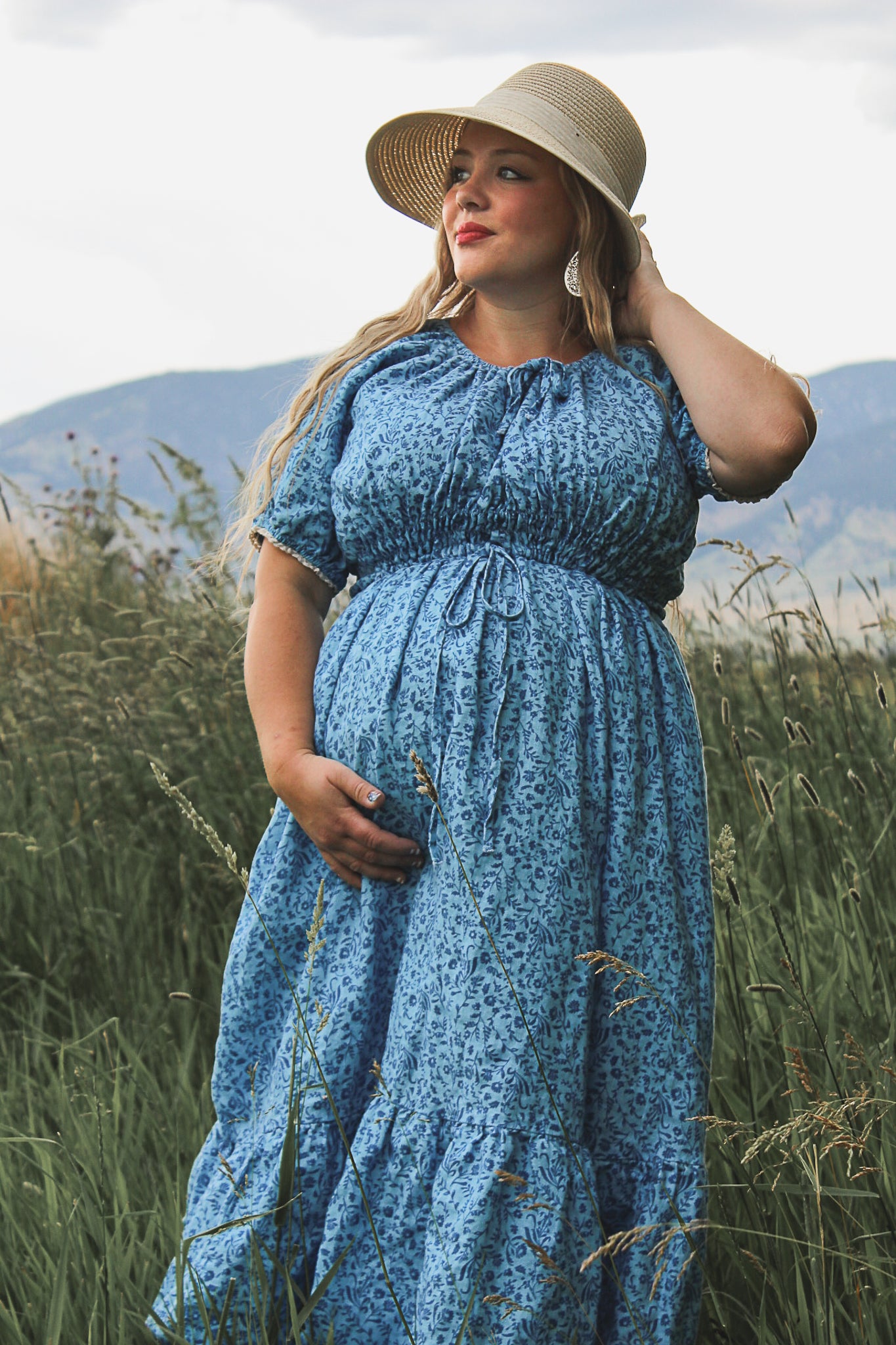 Woman in a blue modest nursing dress and hat standing in a field with mountains in the background