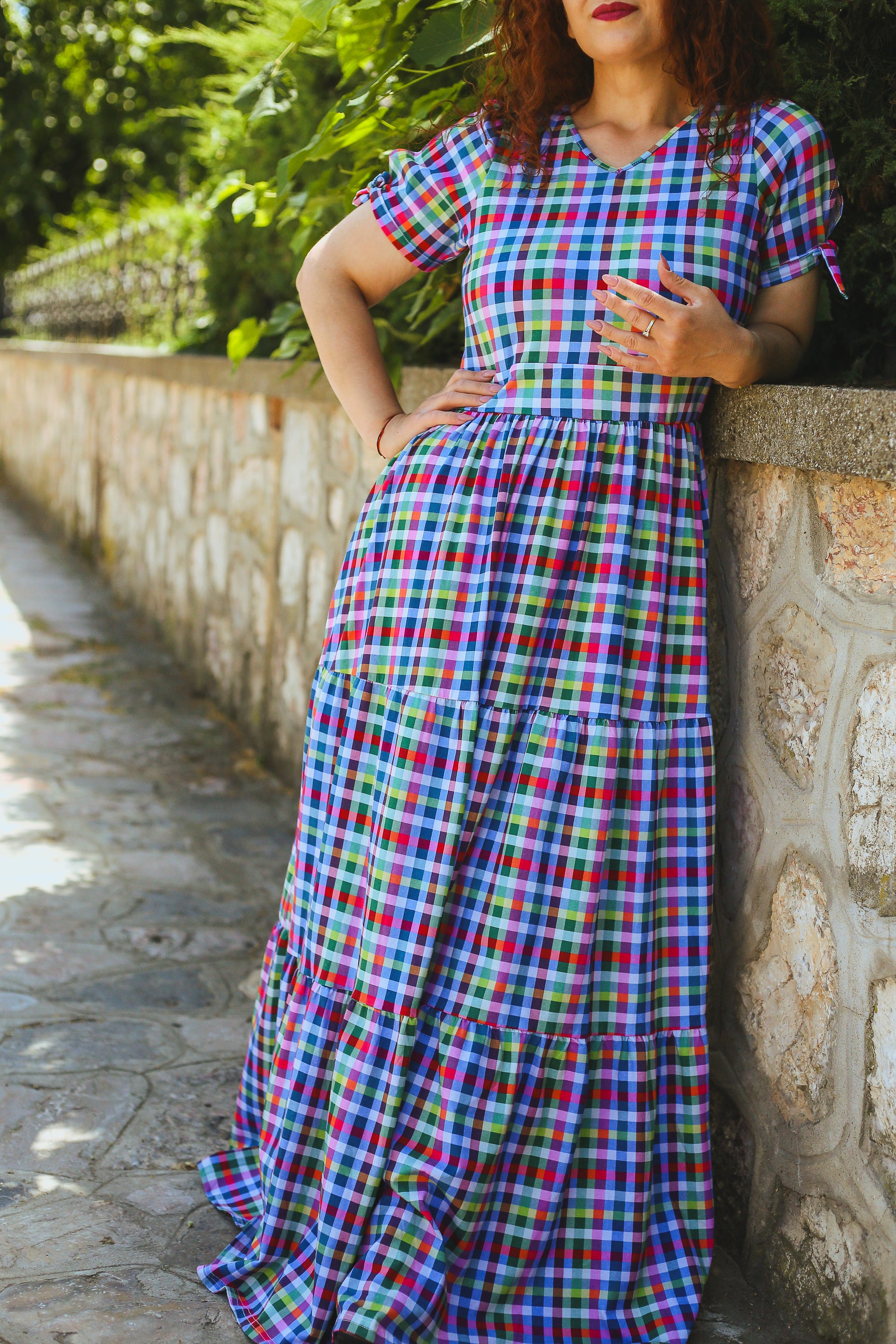 Woman wearing a colorful checkered modest nursing dress leaning against a stone wall with greenery in the background