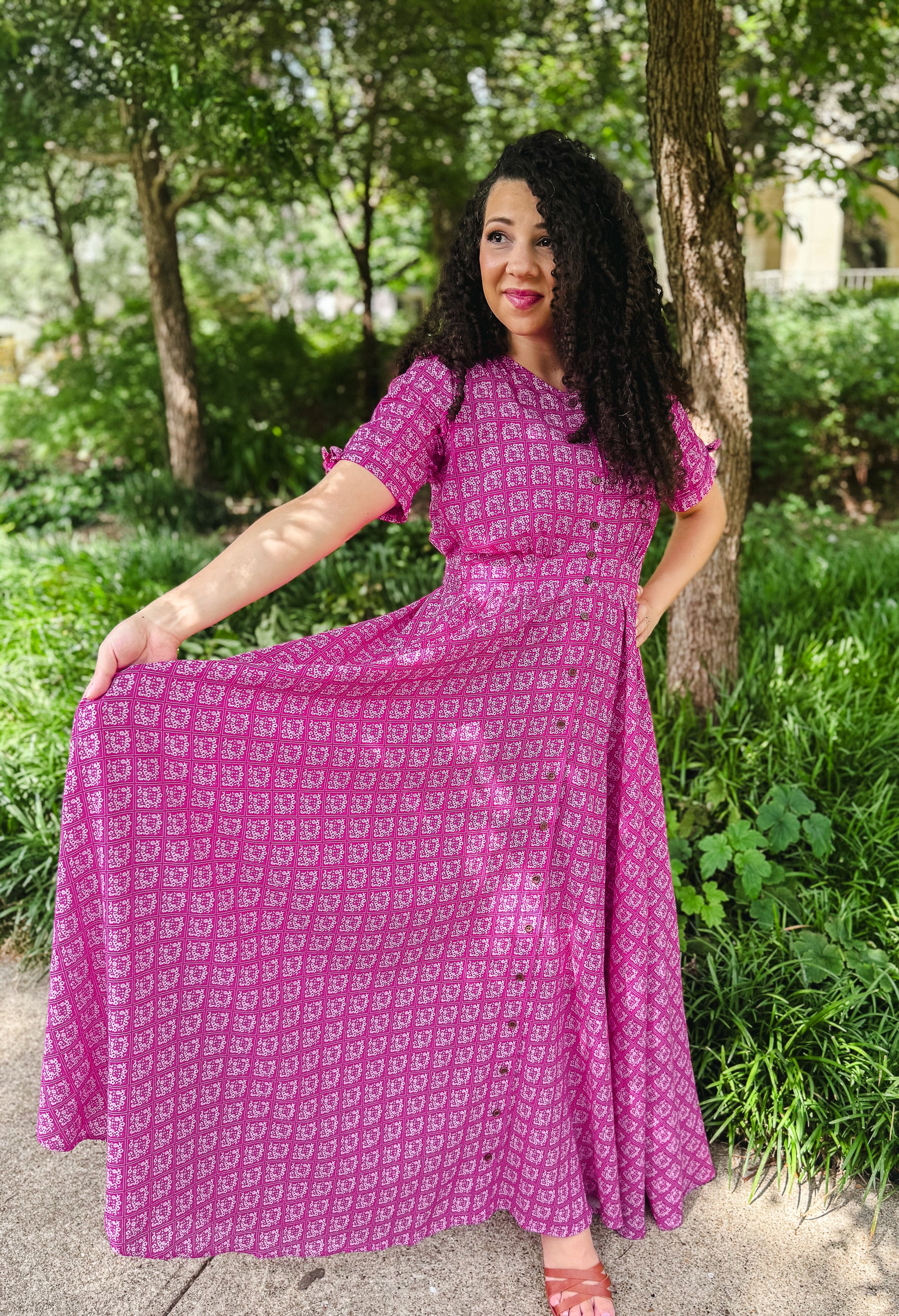 Woman wearing a pink modest nursing dress with a pattern, standing outdoors with trees and greenery in the background.