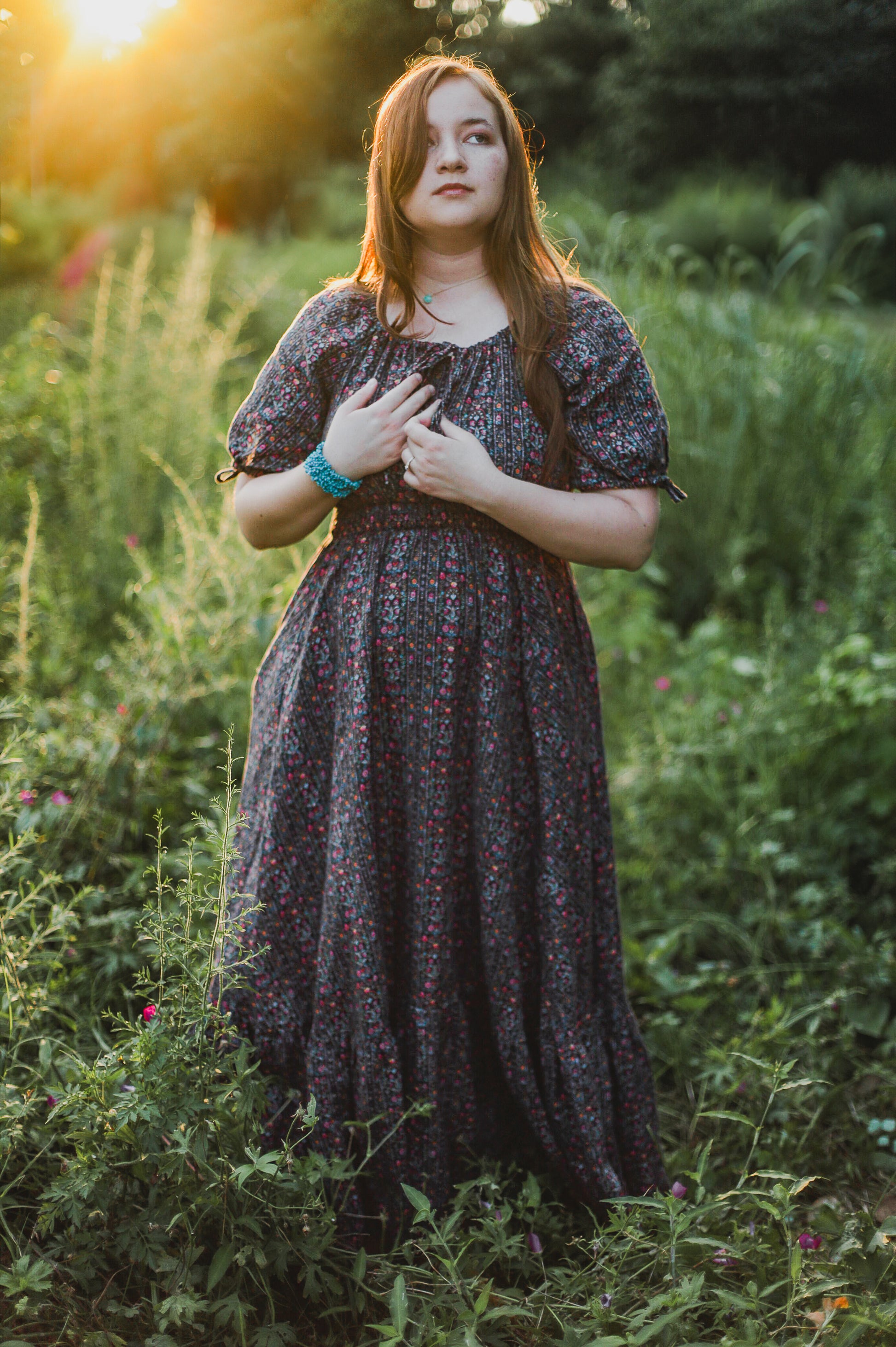 Woman in a floral modest nursing dress standing in a field with sunlight filtering through.