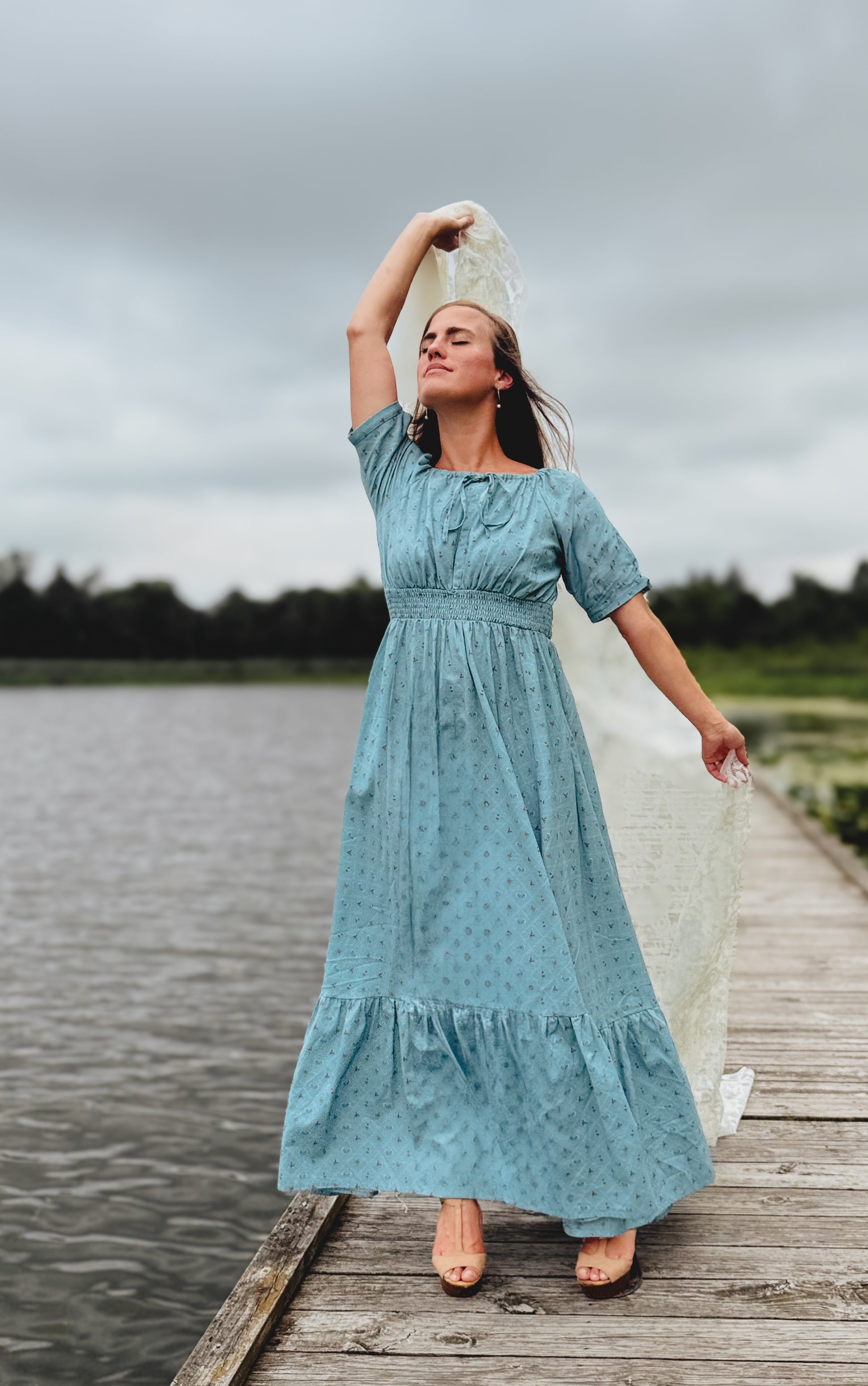 Woman in a teal modest nursing dress standing on a wooden dock by a lake.