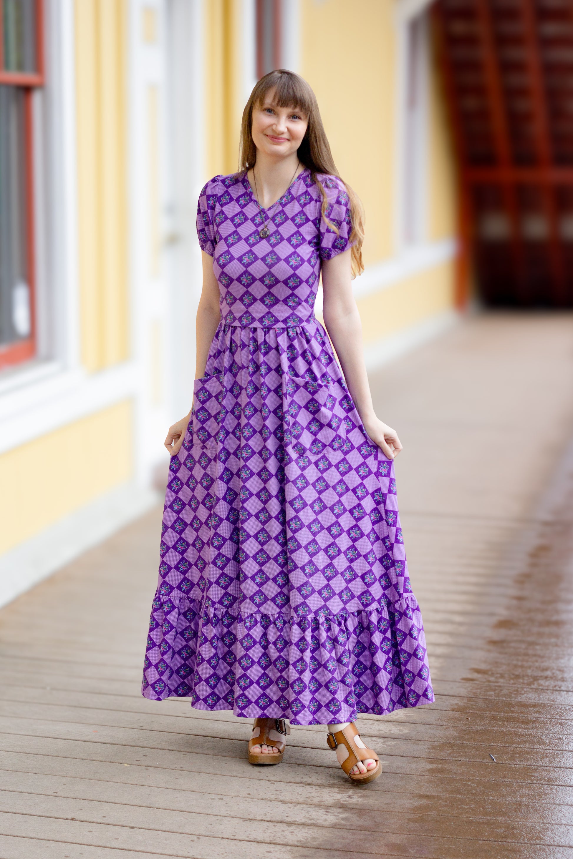 A woman standing on a wooden platform wearing a long purple modest nursing dress with a geometric pattern, featuring a v-neck, elastic sleeve band, natural waistline, ruffle skirt, and elastic back.