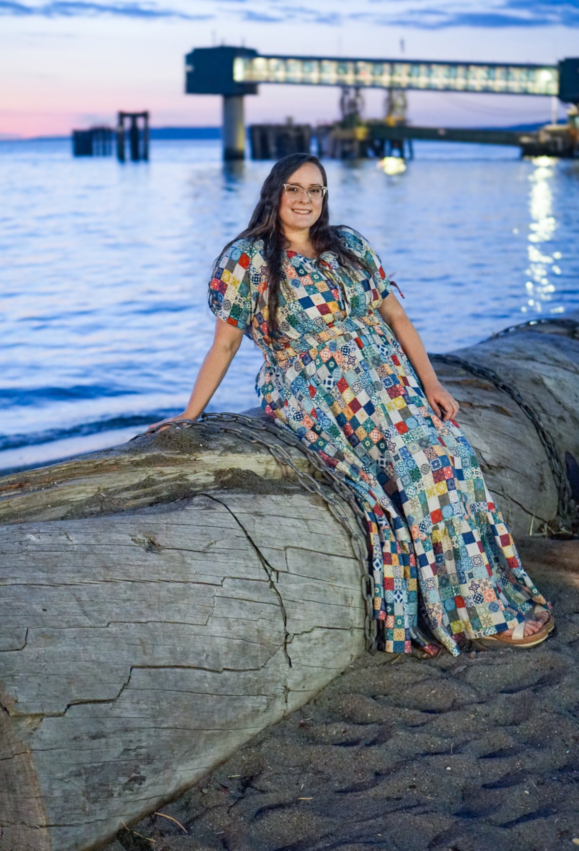 Woman in a colorful modest nursing dress sitting on a log by the water with a bridge in the background