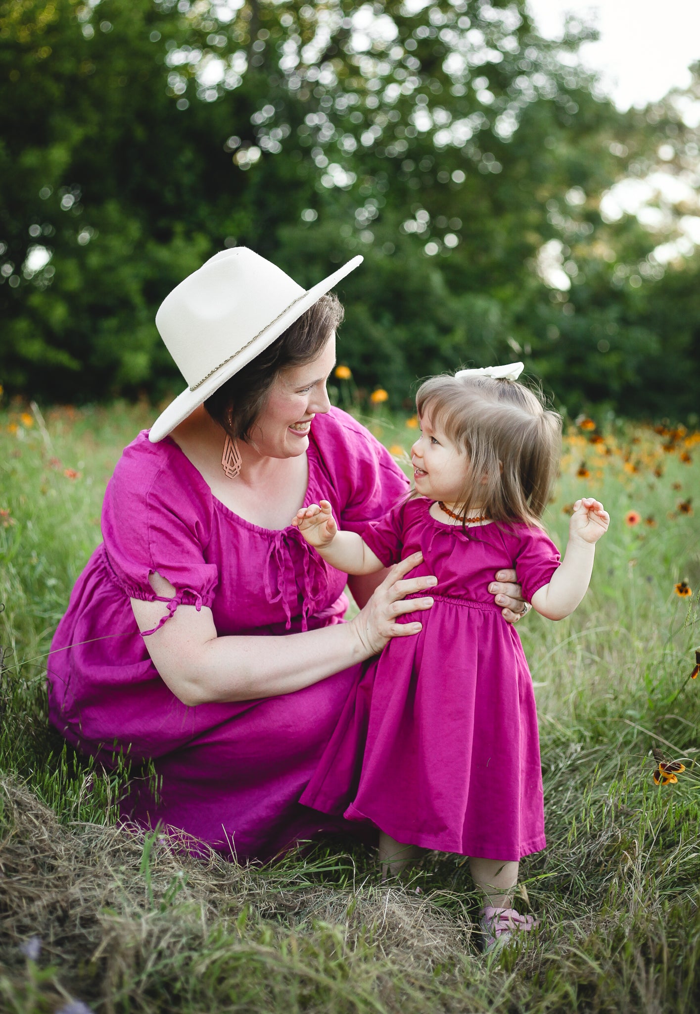 Woman and child in matching pink dresses sitting in a field with greenery and flowers in the background