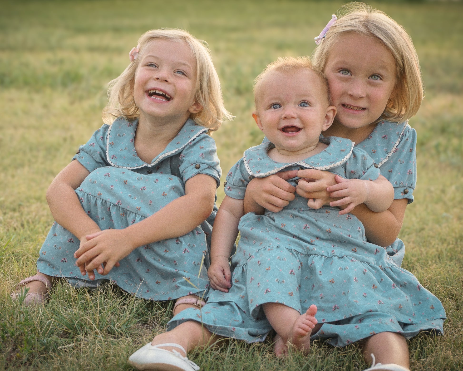 Three children in matching blue dresses sitting on grass