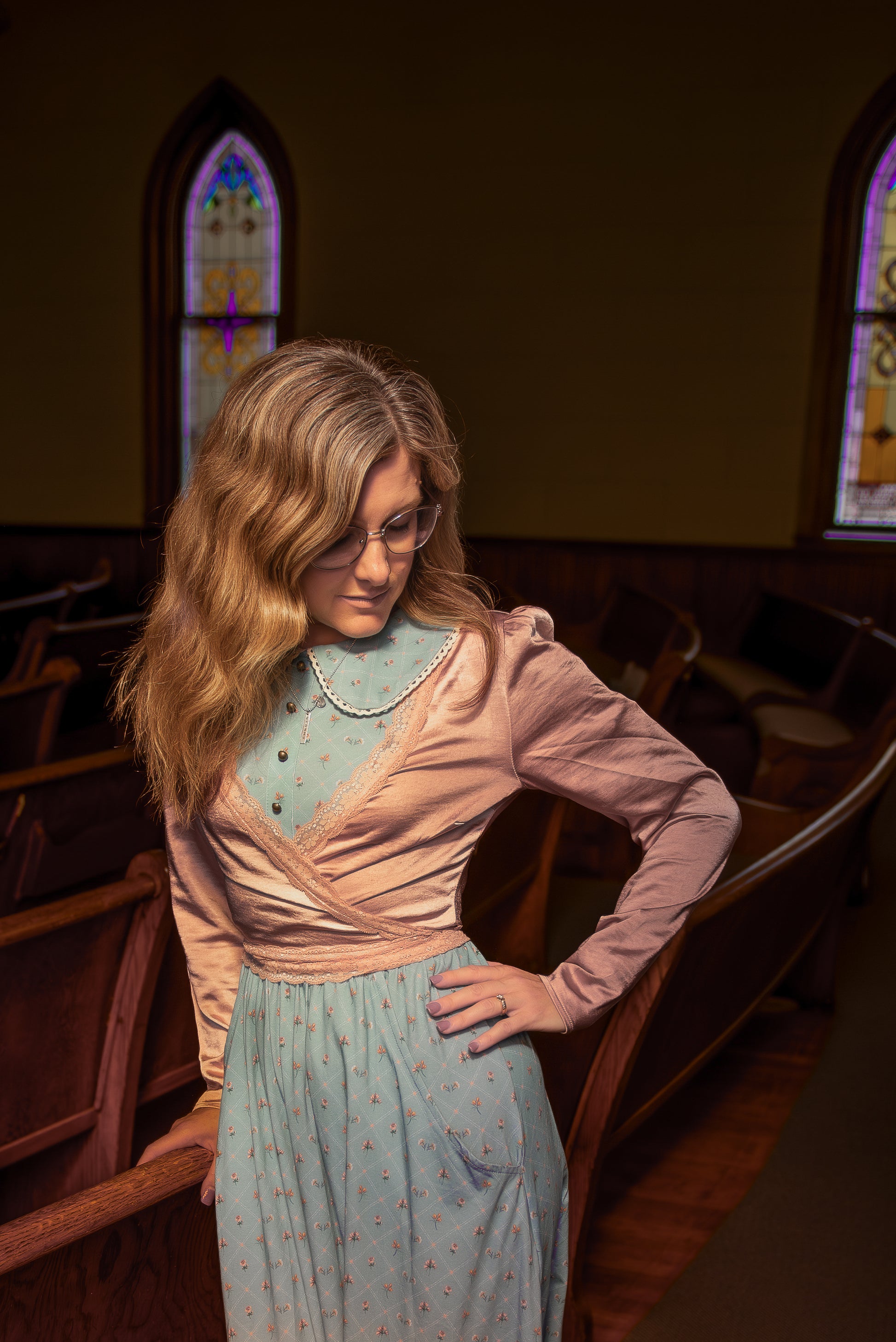 Woman in a vintage-style modest nursing dress standing in a church pew with stained glass windows in the background.
