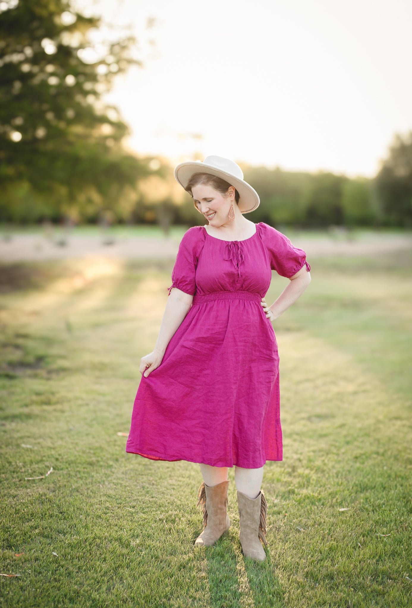 Woman in a pink modest nursing dress and white hat standing in a park with trees and grass in the background.
