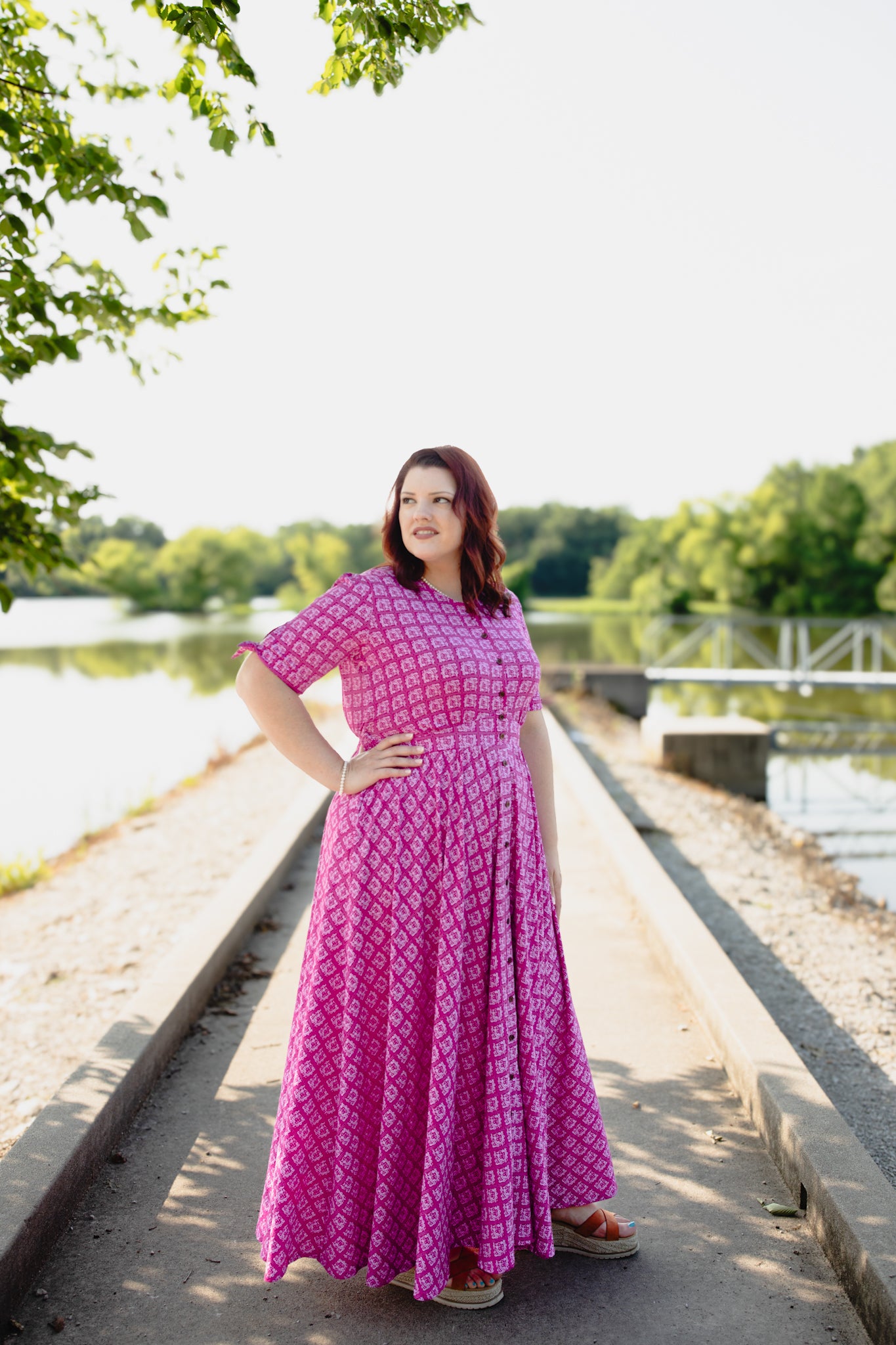 Woman in a pink modest nursing dress standing by a river with trees in the background