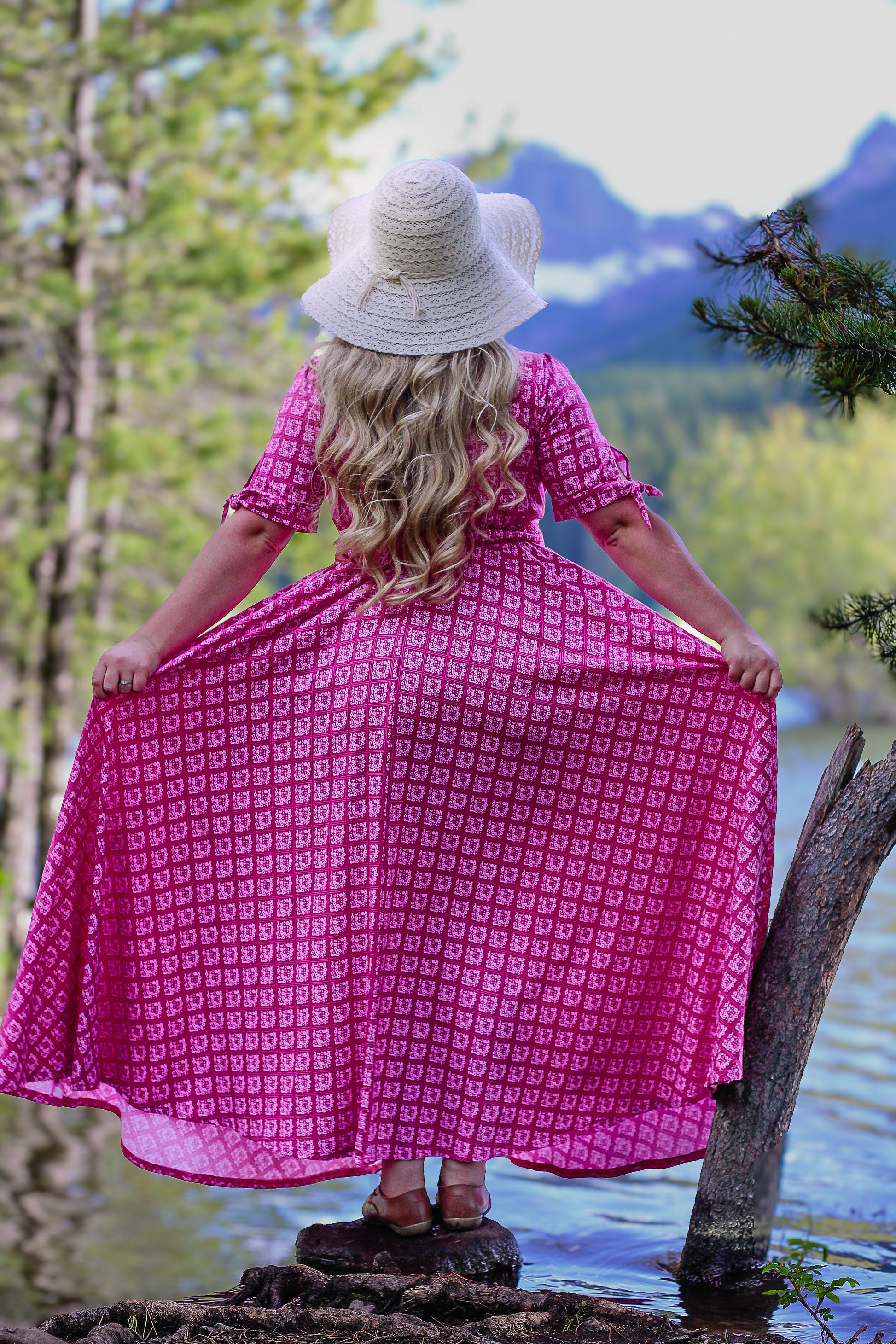 Woman in a pink modest nursing dress standing by a lake with mountains in the background