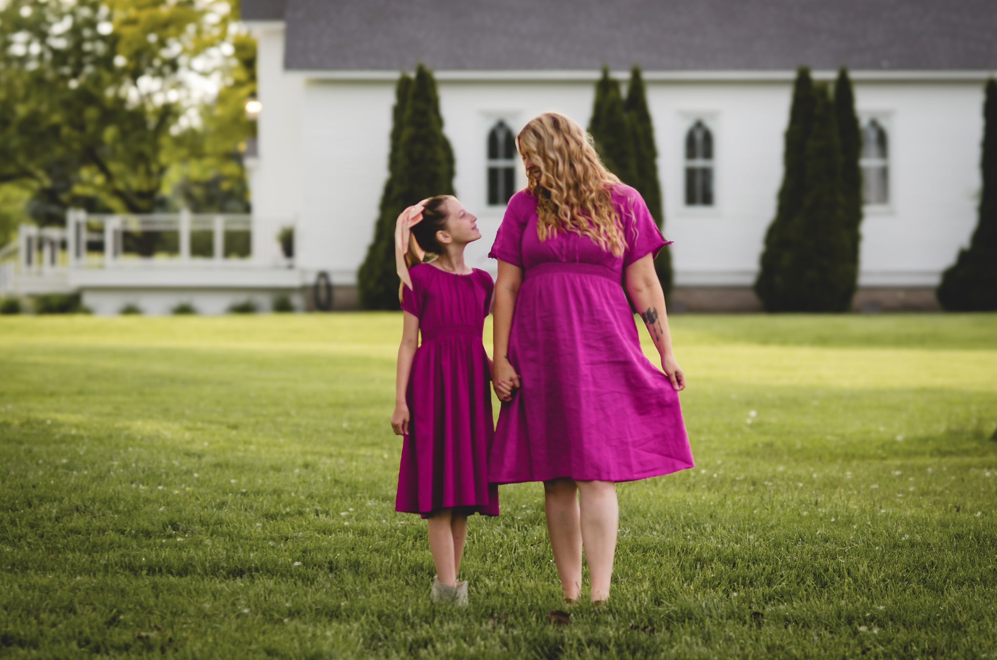 Mother and daughter in matching pink modest dresses standing on a grassy field with a white building in the background.