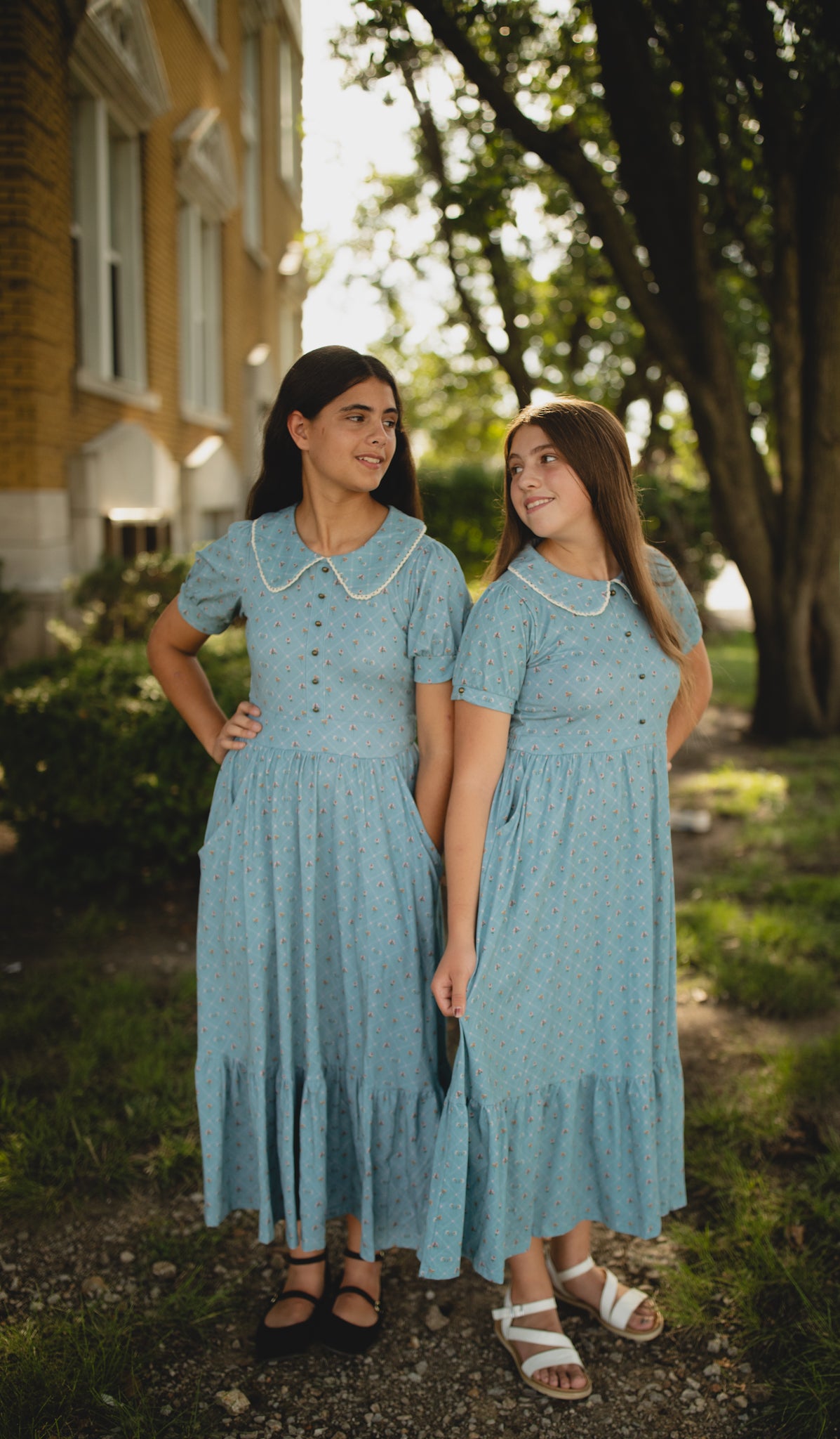 Two women in matching blue modest dresses standing outdoors with trees and a building in the background.