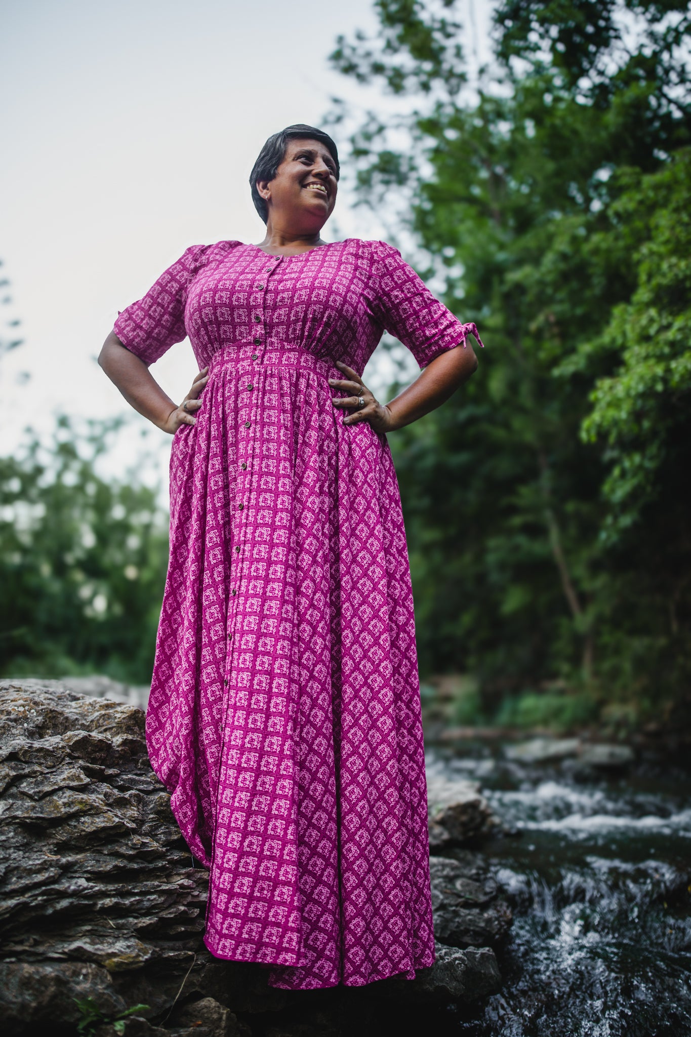 Woman in a pink modest nursing dress standing by a stream with trees in the background