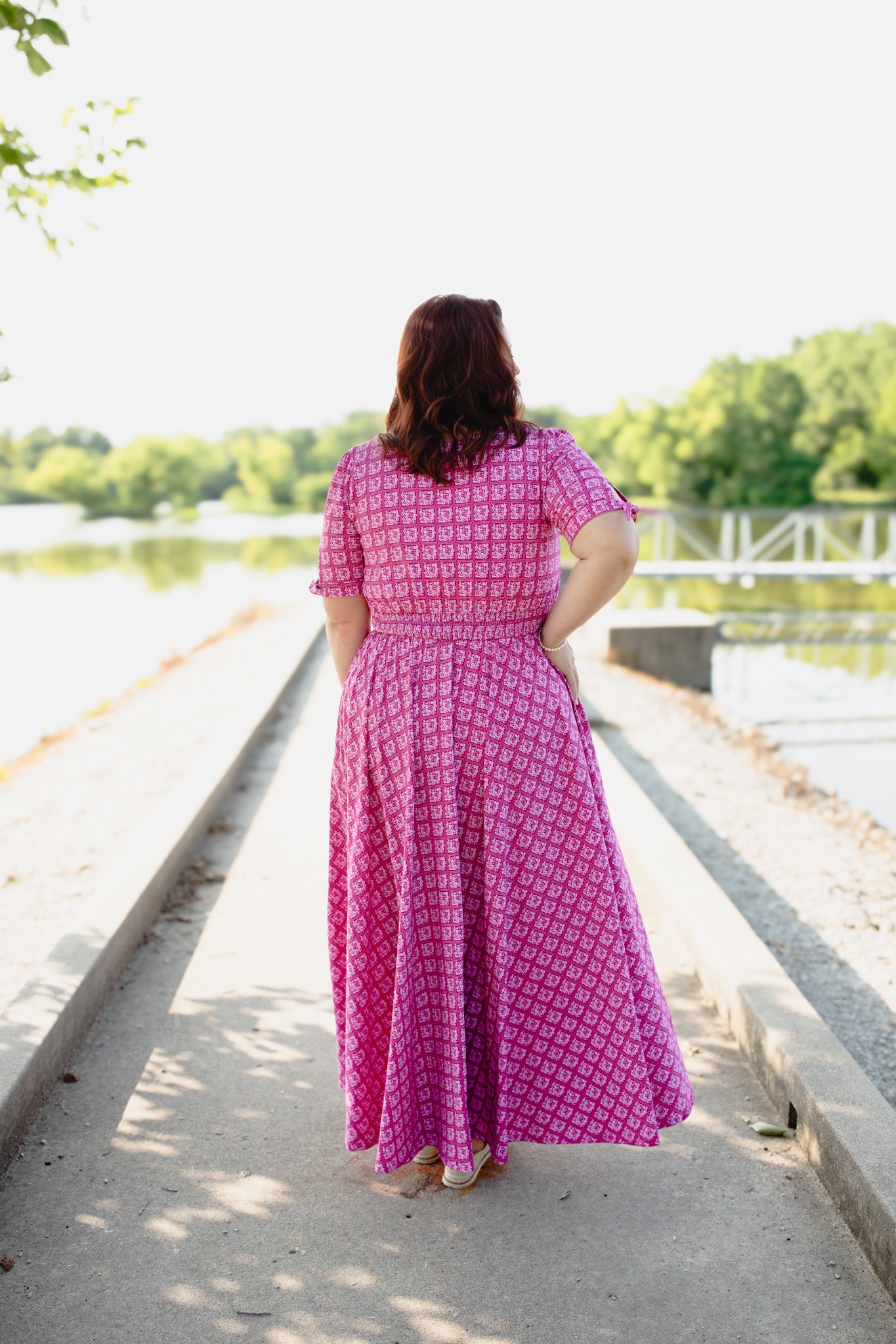 Woman in a pink modest nursing dress walking along a path by a lake with trees in the background