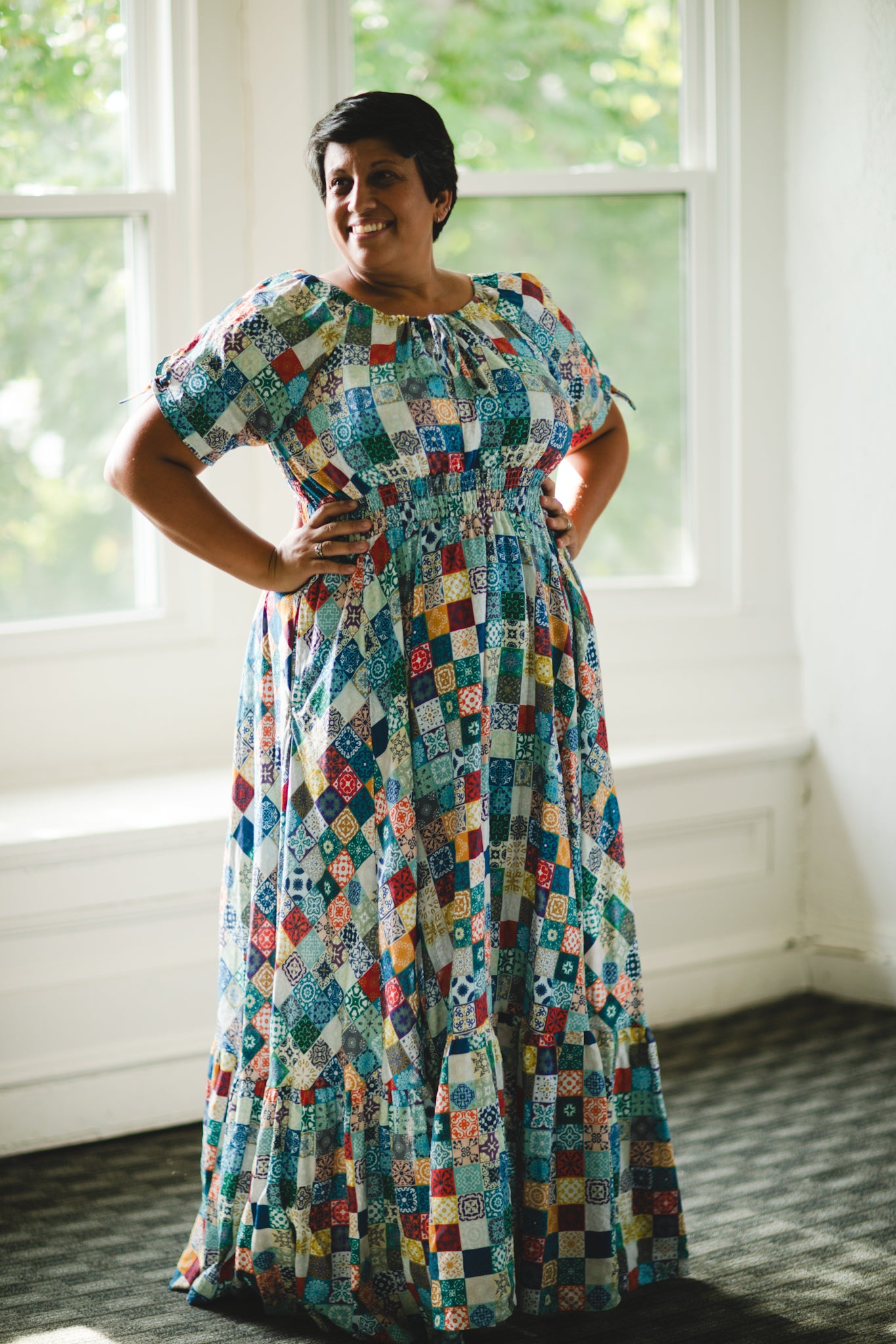 Woman wearing a colorful patterned modest nursing dress standing in a room with large windows.