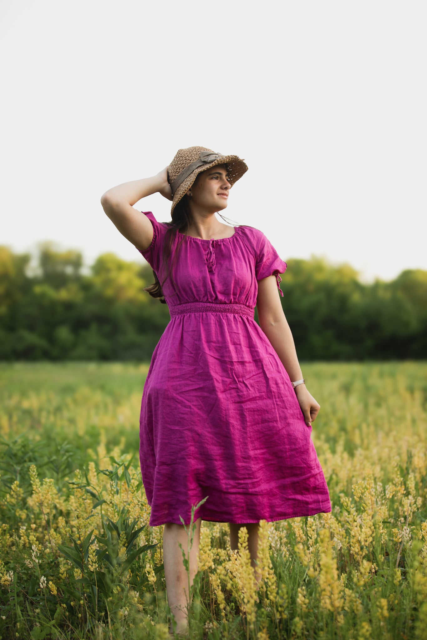 Woman in a purple modest nursing dress and straw hat standing in a field with yellow flowers.