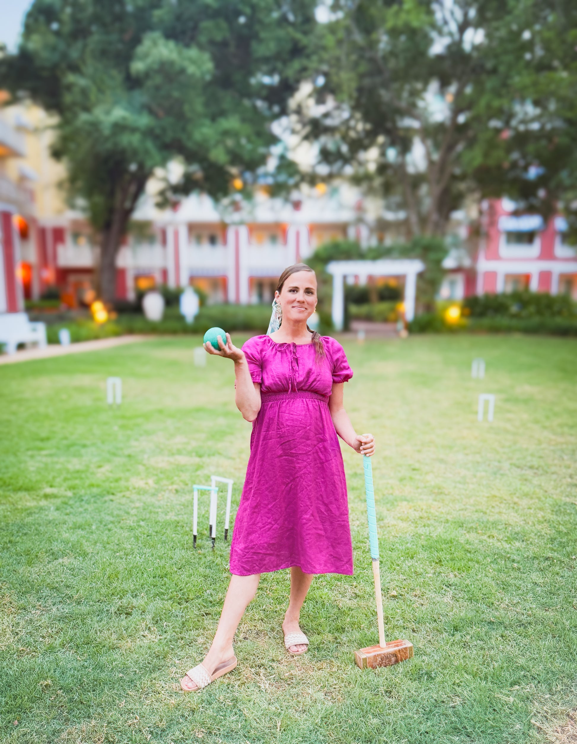 Woman in a pink modest nursing dress holding a croquet mallet and ball on a grassy lawn with a building in the background.