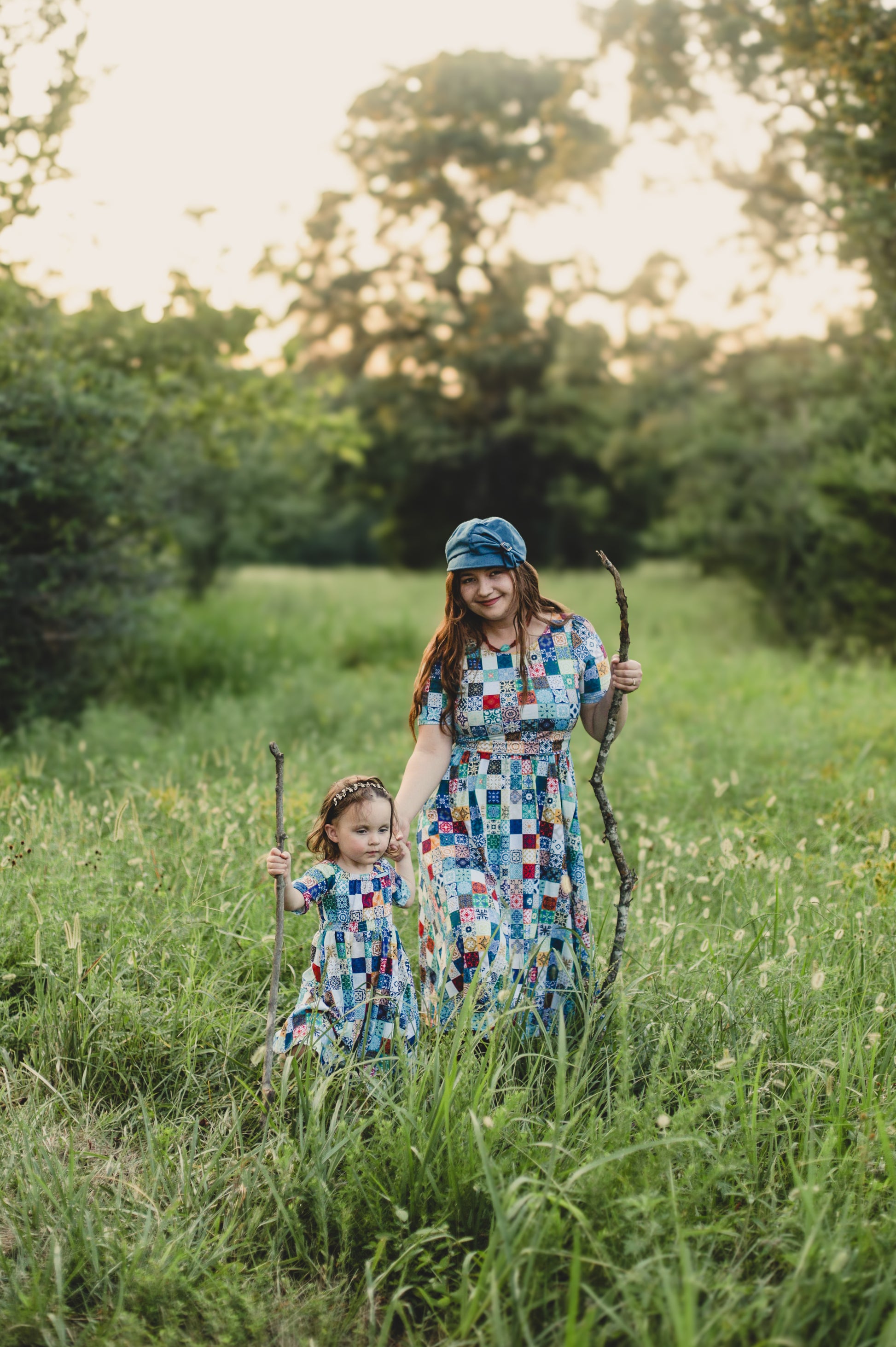 Mother and child in colorful checkered modest dresses holding sticks in a grassy field with trees in the background.