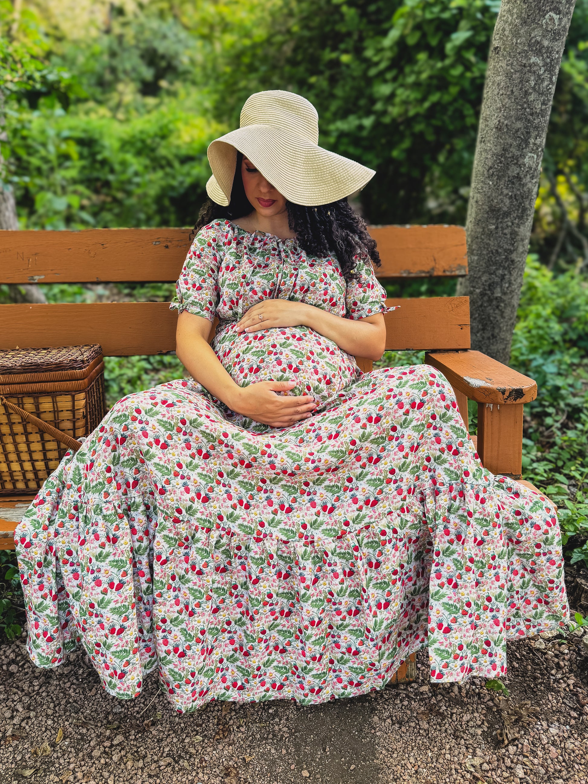 Woman in a floral modest maternity dress and sun hat sitting on a bench in a garden.