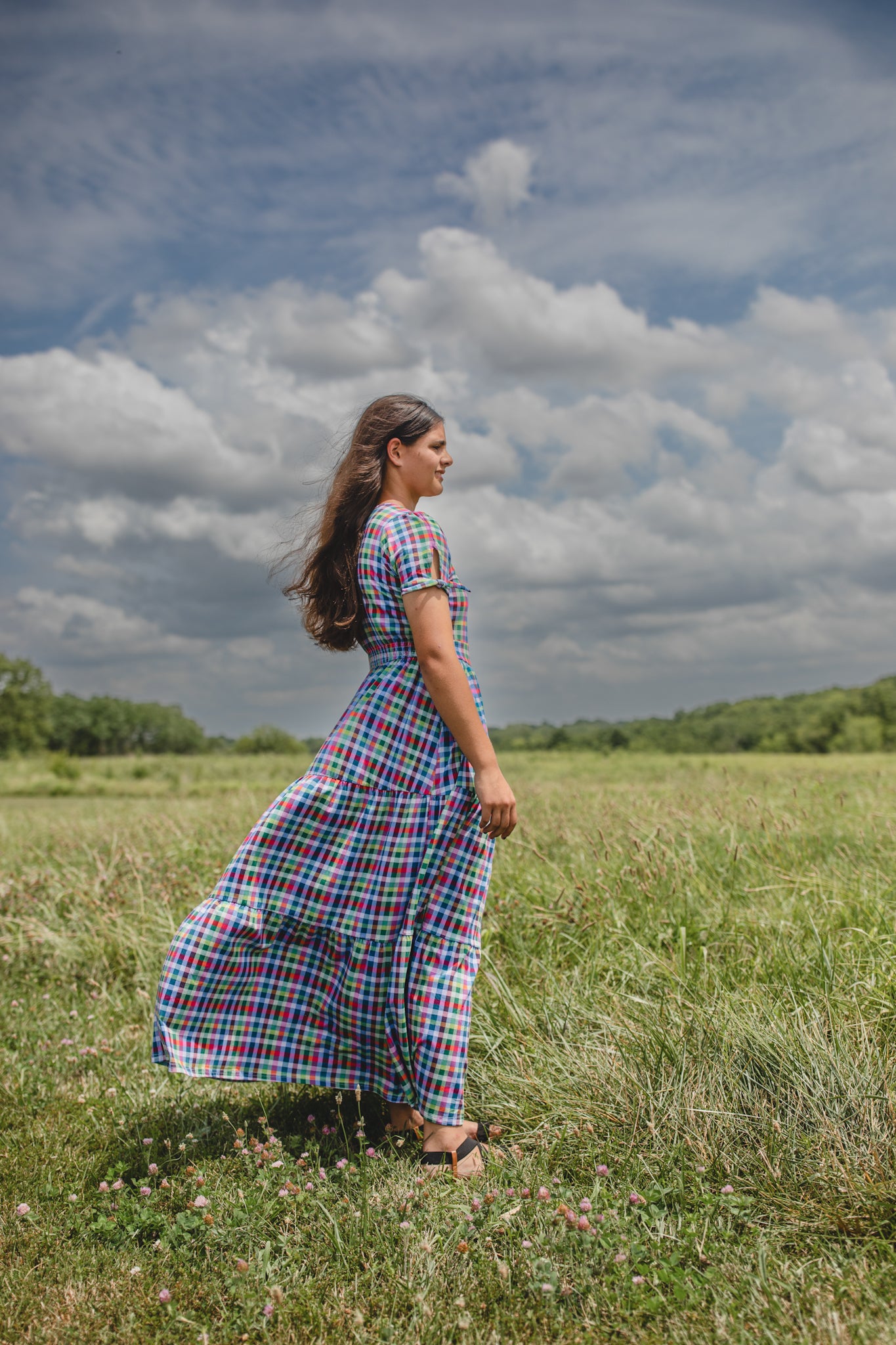Woman in a plaid modest nursing dress standing in a grassy field with a cloudy sky.