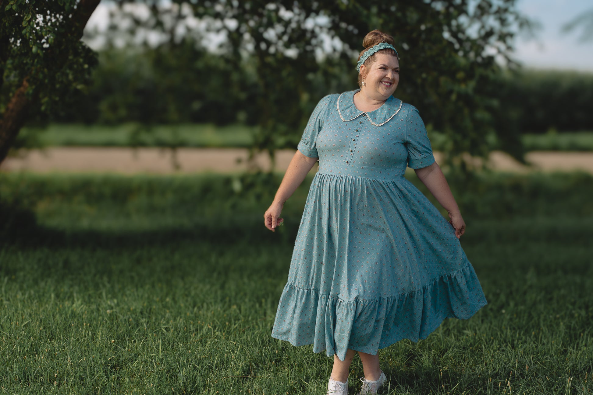 Woman in a blue modest nursing dress standing in a grassy field with trees in the background