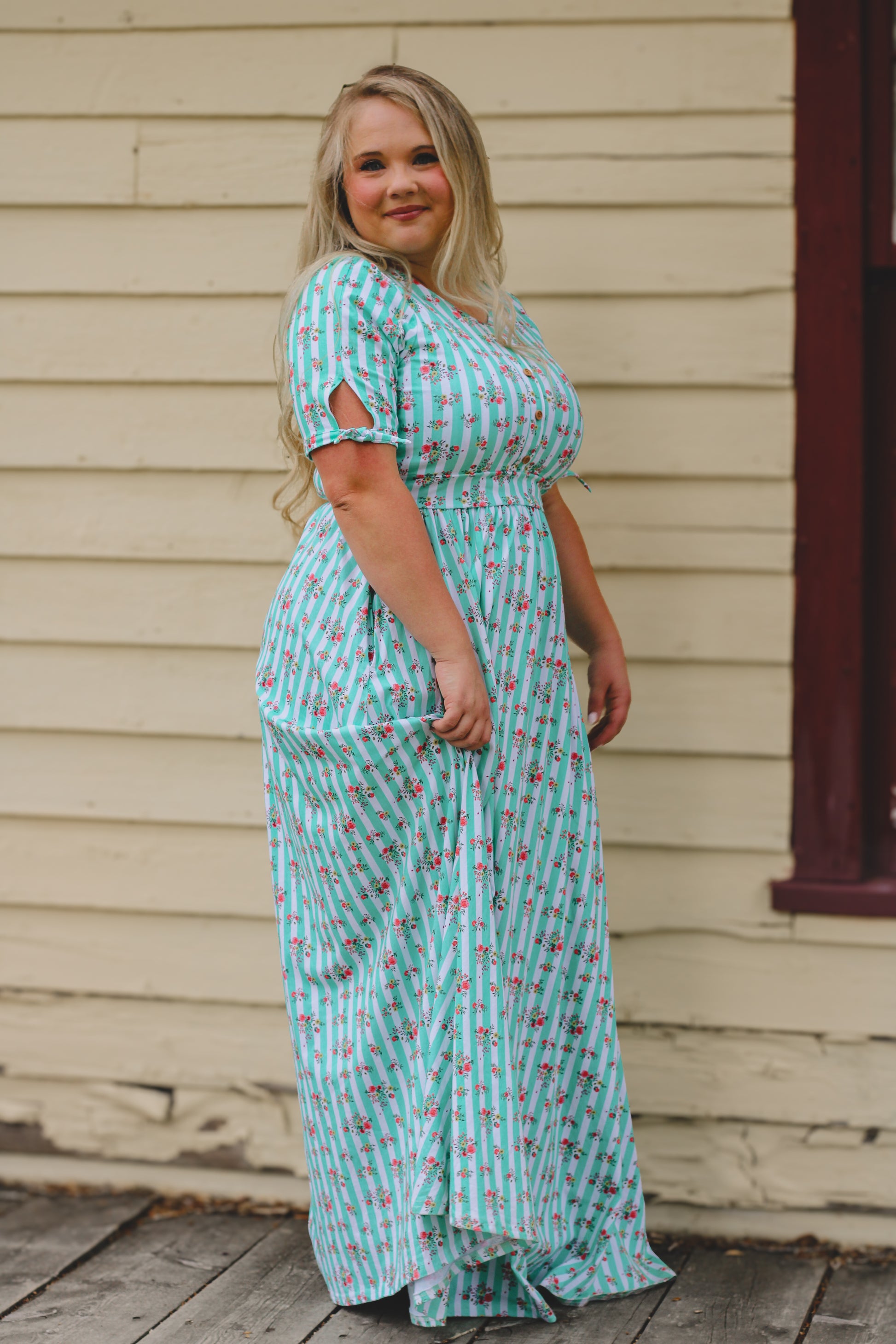 Woman wearing a turquoise floral modest nursing dress standing in front of a beige wall.