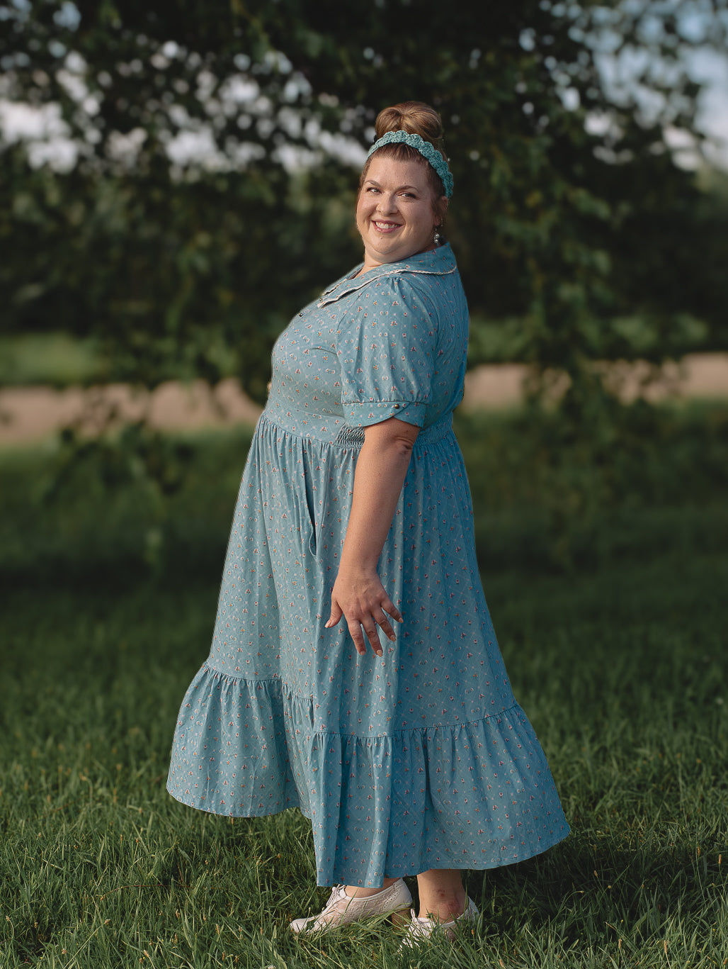 Woman in a blue modest nursing dress standing in a grassy field with trees in the background