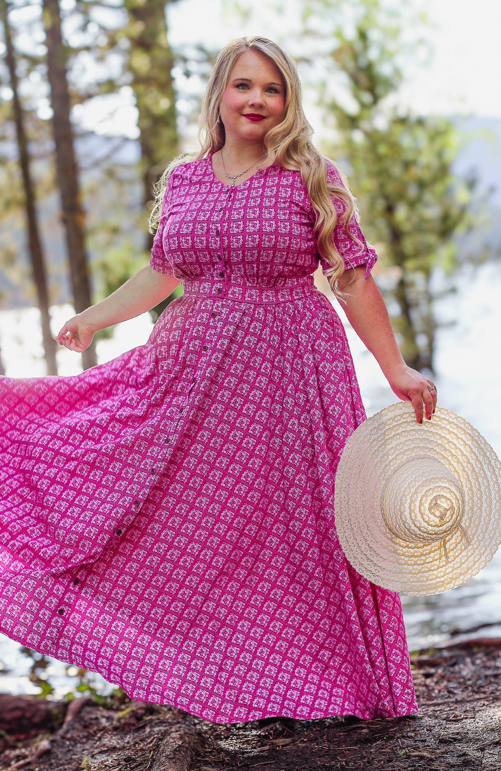Woman in a pink modest nursing dress holding a straw hat outdoors with trees and water in the background