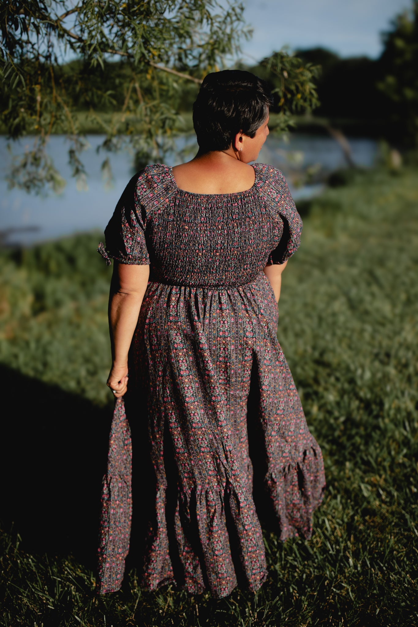 Person wearing a patterned modest nursing dress standing in a natural setting with trees and water.