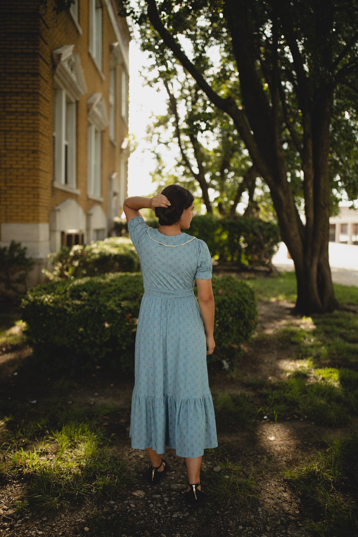 Woman in a blue modest dress standing outdoors near a tree and building