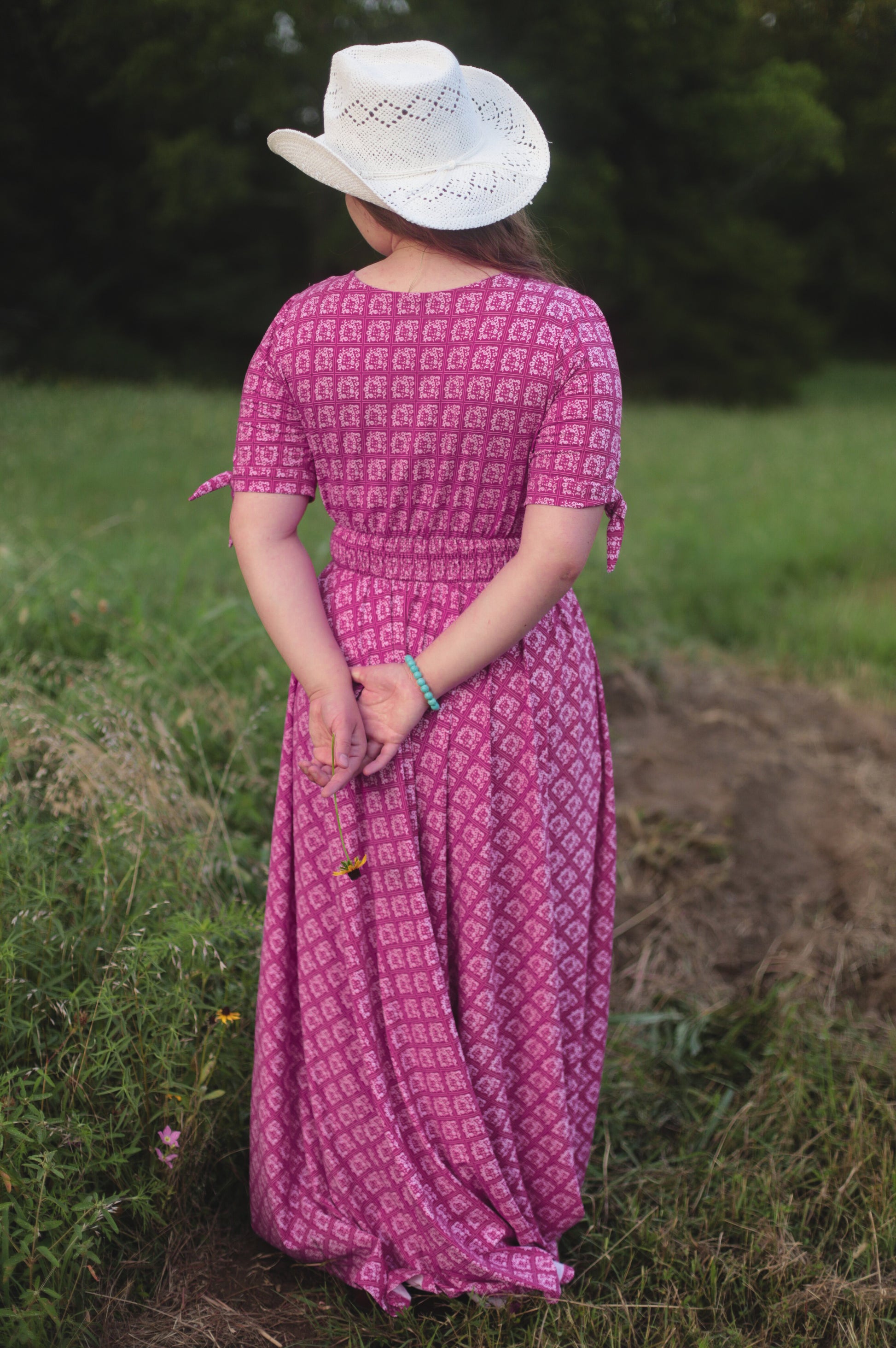 Person wearing a pink modest nursing dress and white cowboy hat standing in a grassy field.
