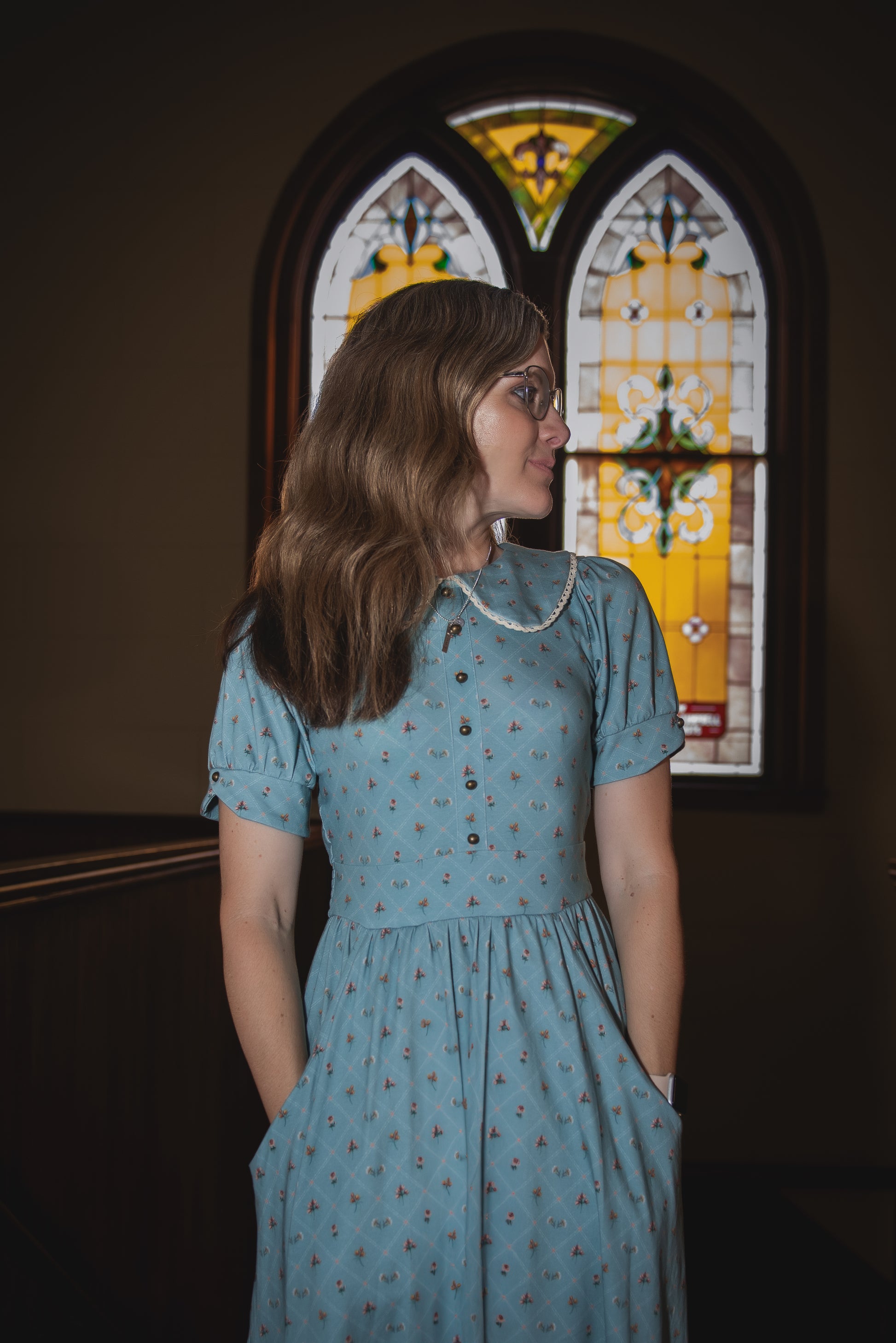Woman in a blue modest nursing dress standing in front of a stained glass window