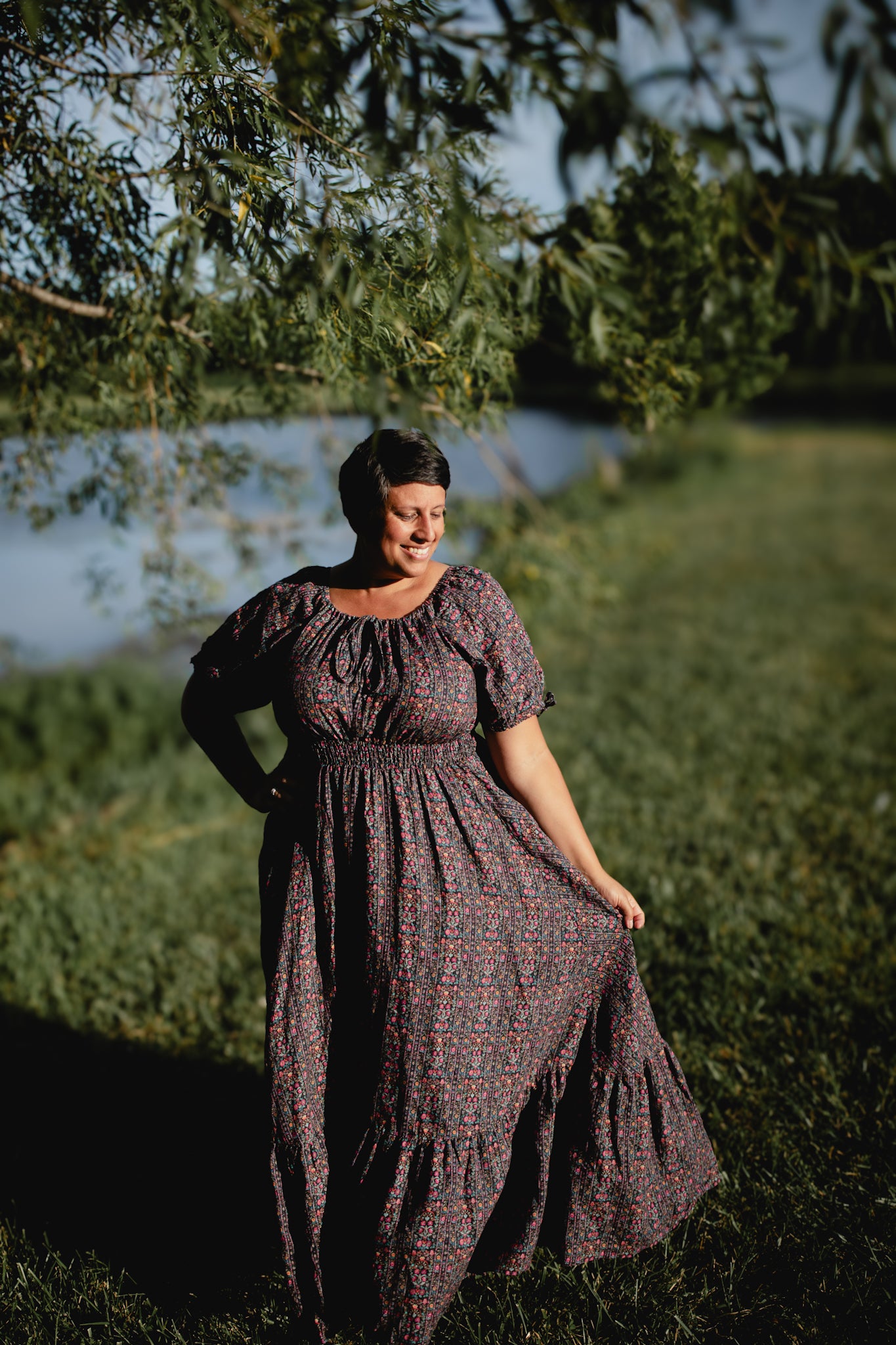 Woman in a long, dark floral modest nursing dress standing in a grassy area with trees and water in the background.