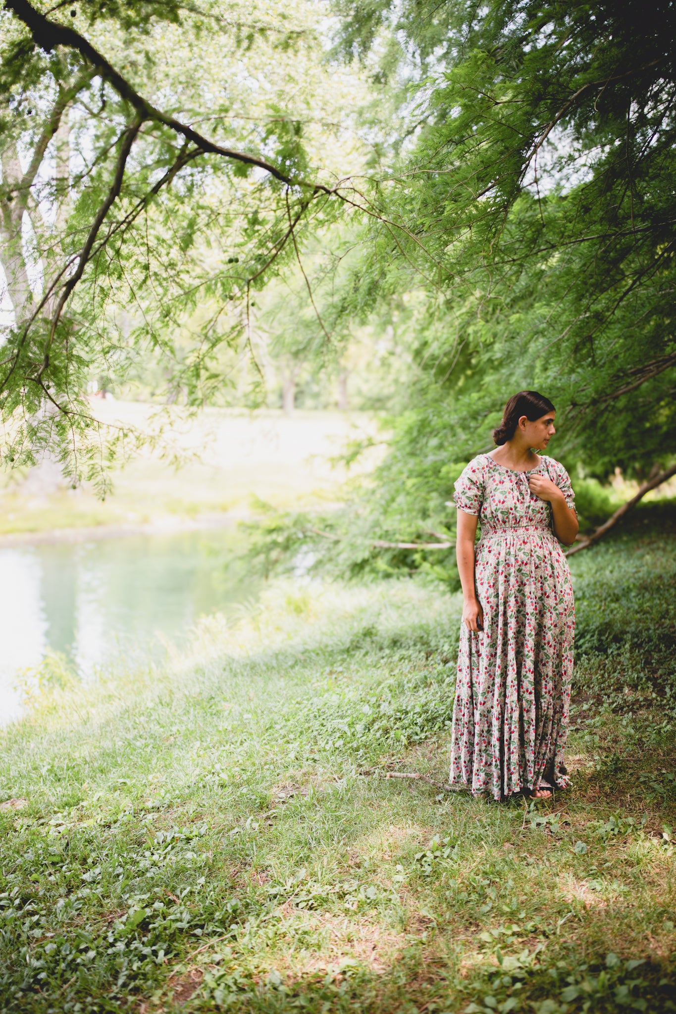 Woman in a floral modest nursing dress standing in a park with trees and a pond in the background