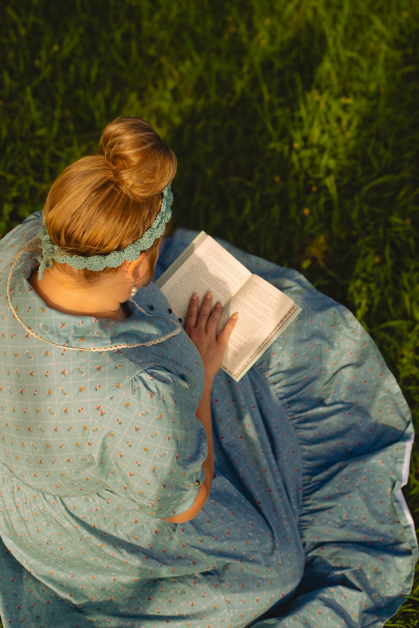 Woman in a blue modest nursing dress sitting on grass reading a book