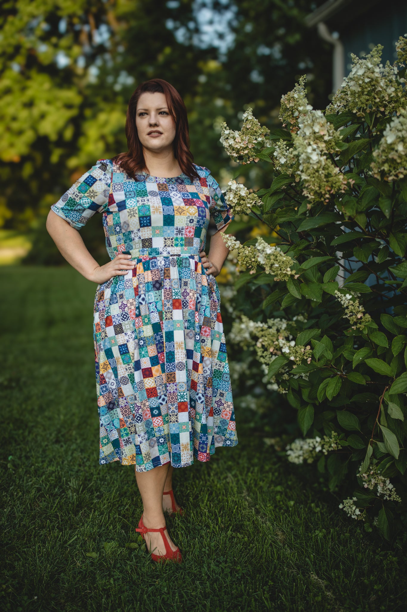 Woman wearing a colorful patchwork modest nursing dress standing outdoors near greenery.