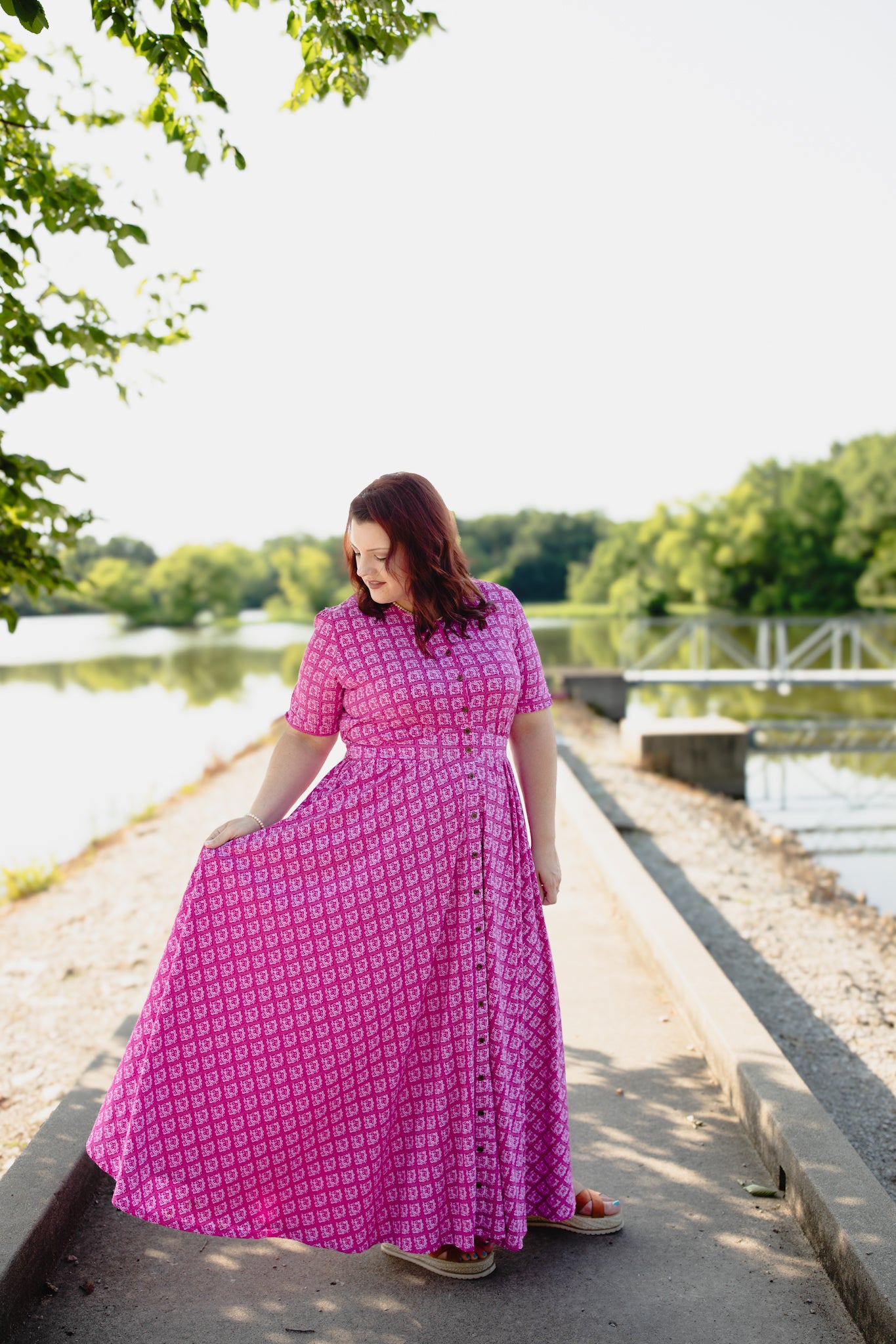Woman in a pink modest nursing dress standing by a lake with trees in the background