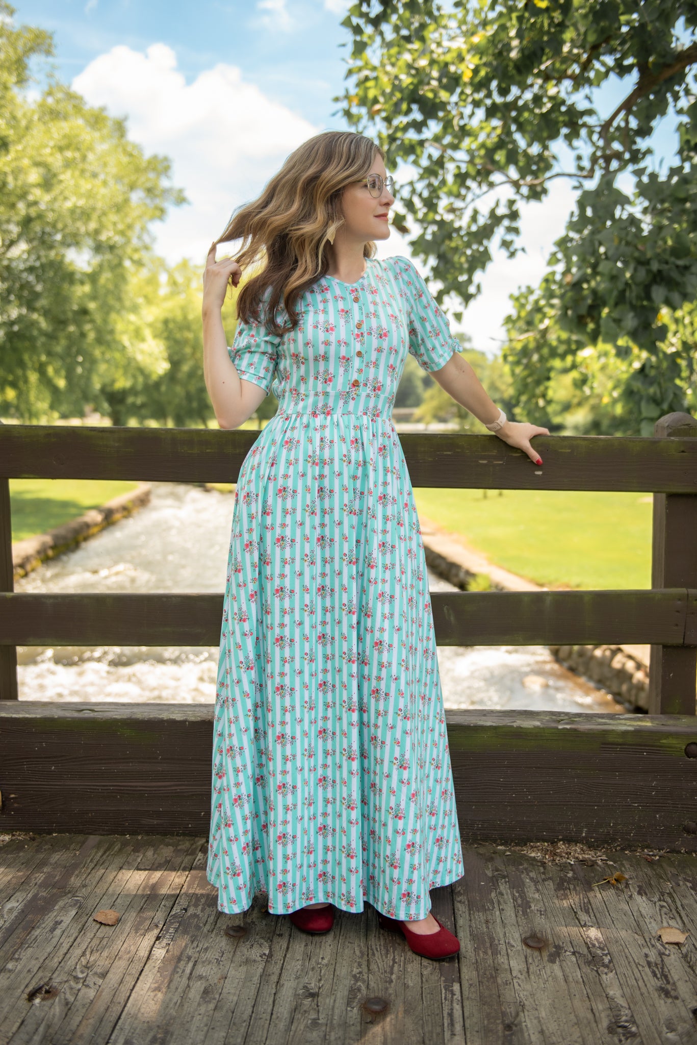 Woman in a long modest nursing dress standing on a wooden bridge with greenery in the background